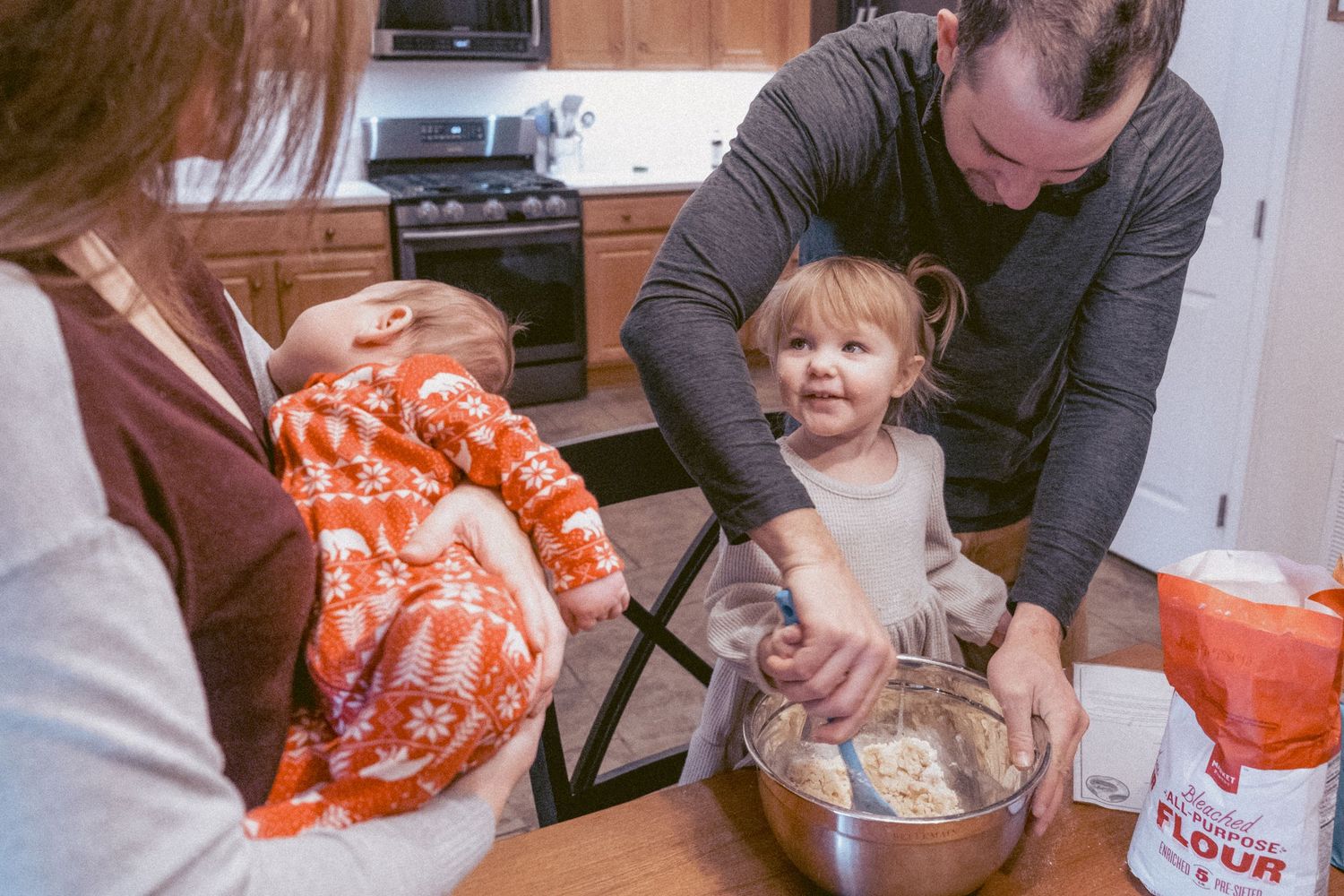 Documentary portrait of child and father mixing Christmas cookies while mother holds newborn, wearing red Christmas pajamas.