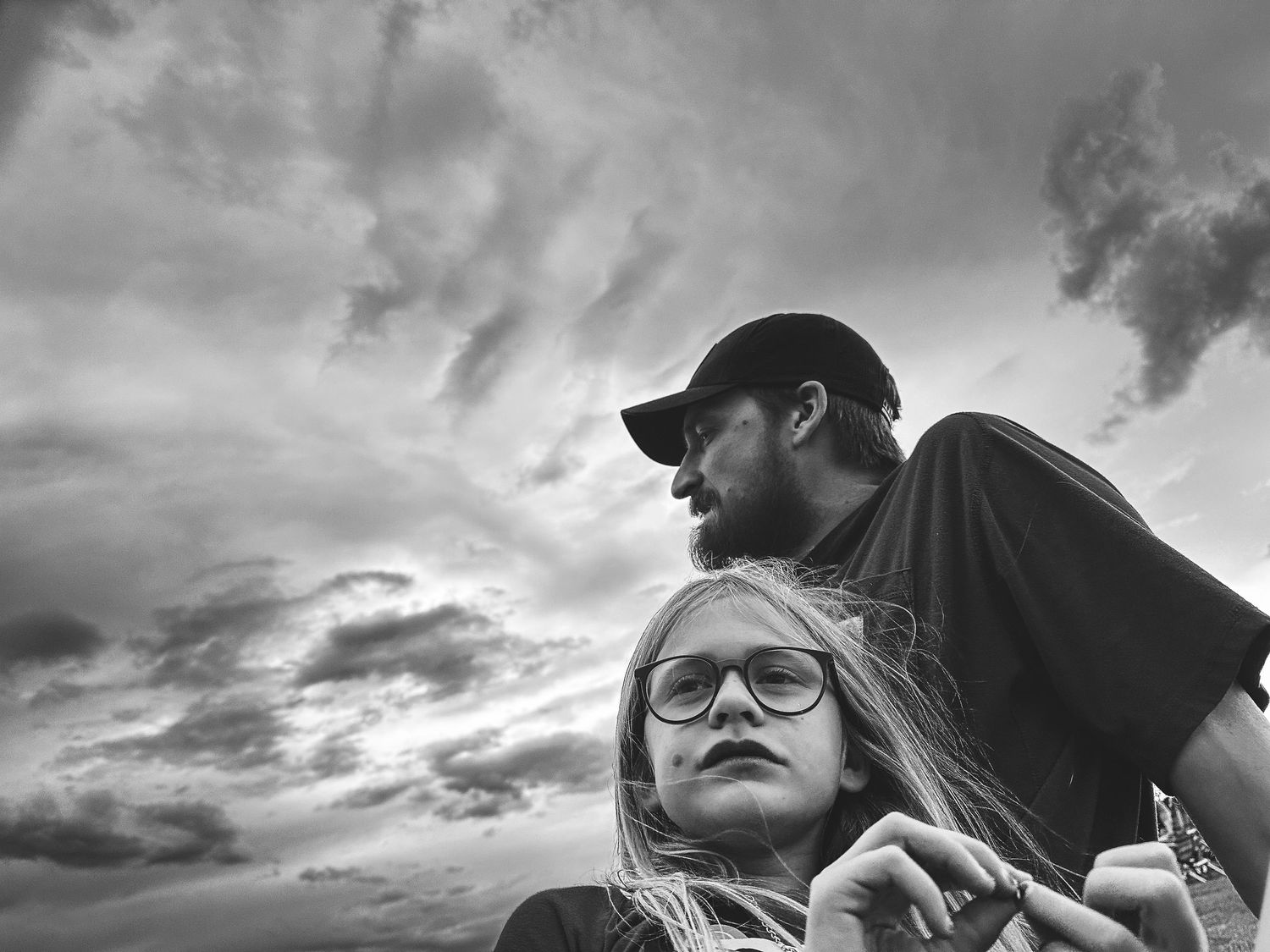 Black and white portrait of girl sitting with father, dramatic clouds in background.