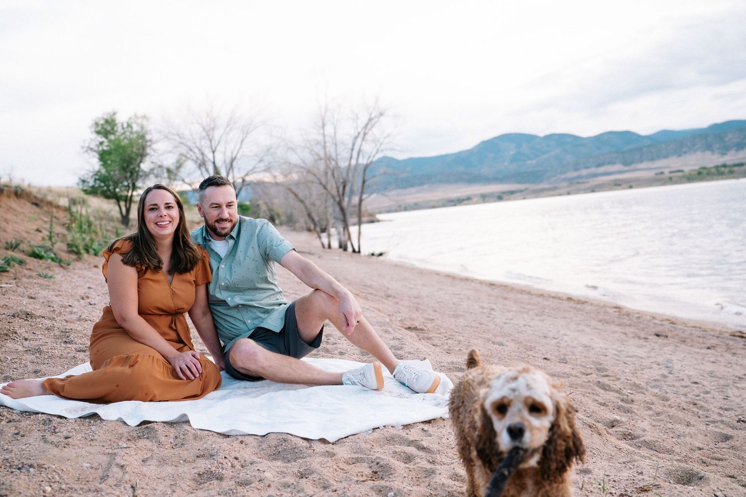 Husband and wife sitting on white blanket at sandy area of Chatfield state park, dog walking toward camera.