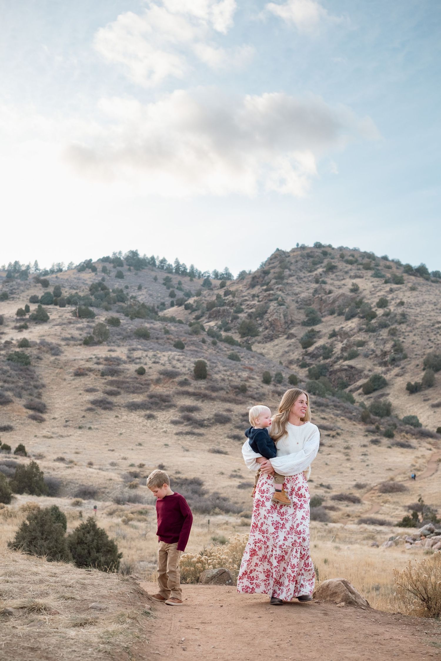 Mother holding one year old while five year old stands nearby.  Foothills views in background, with blue skies and fluffy clouds.