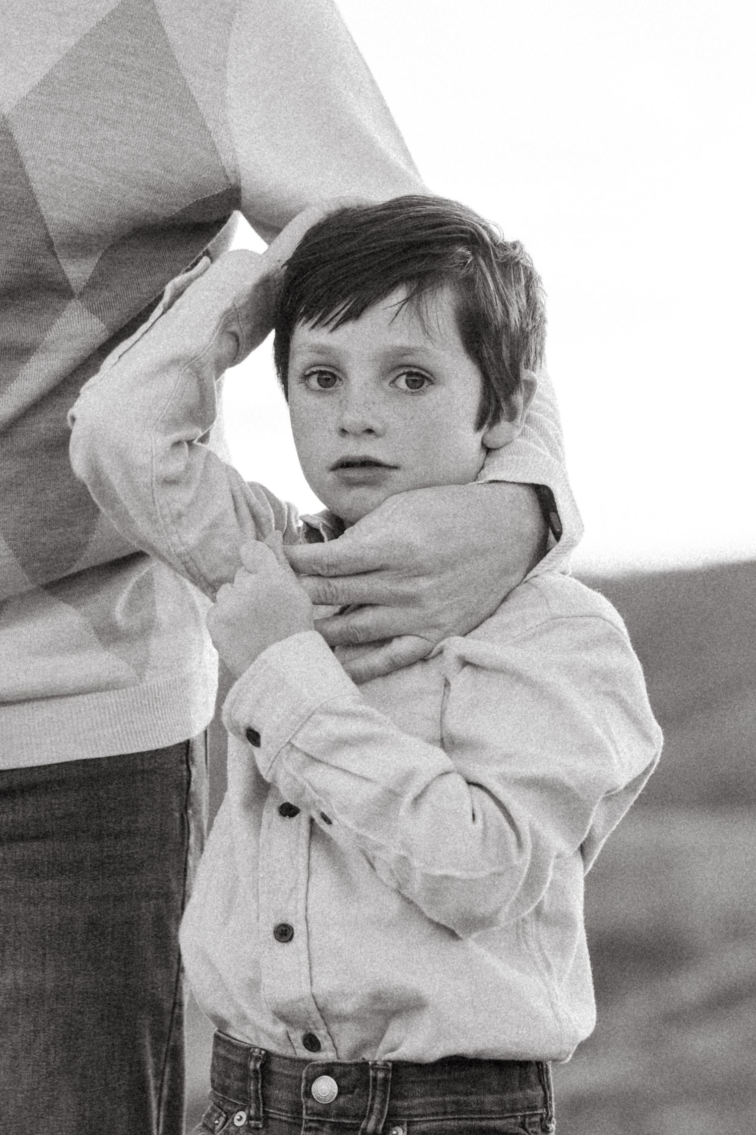 Balck and white portrait of young boy with serious expression, father's hand wrapped around him lovingly.