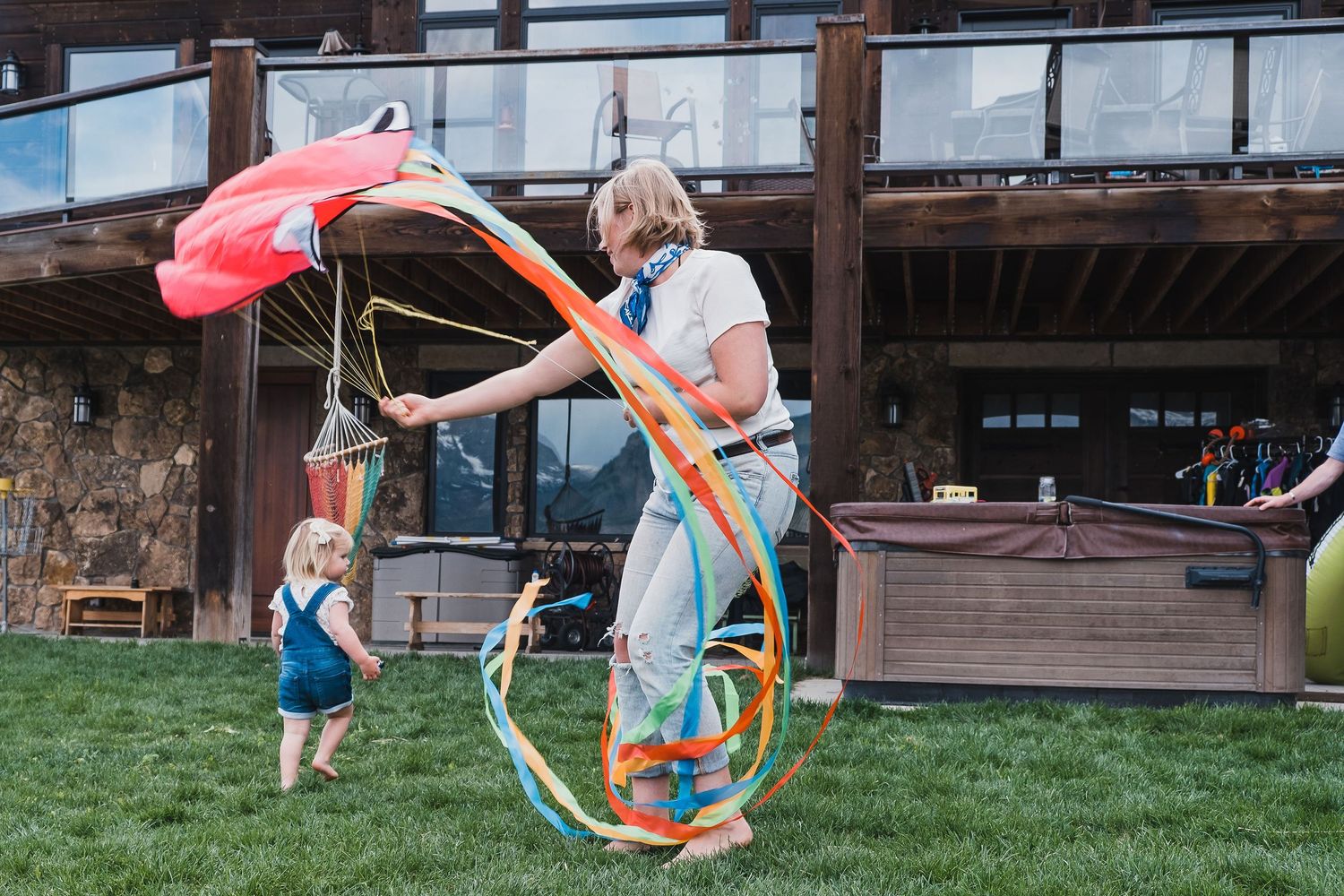 Aunt playing with a colorful kite with toddler niece in front of cabin.