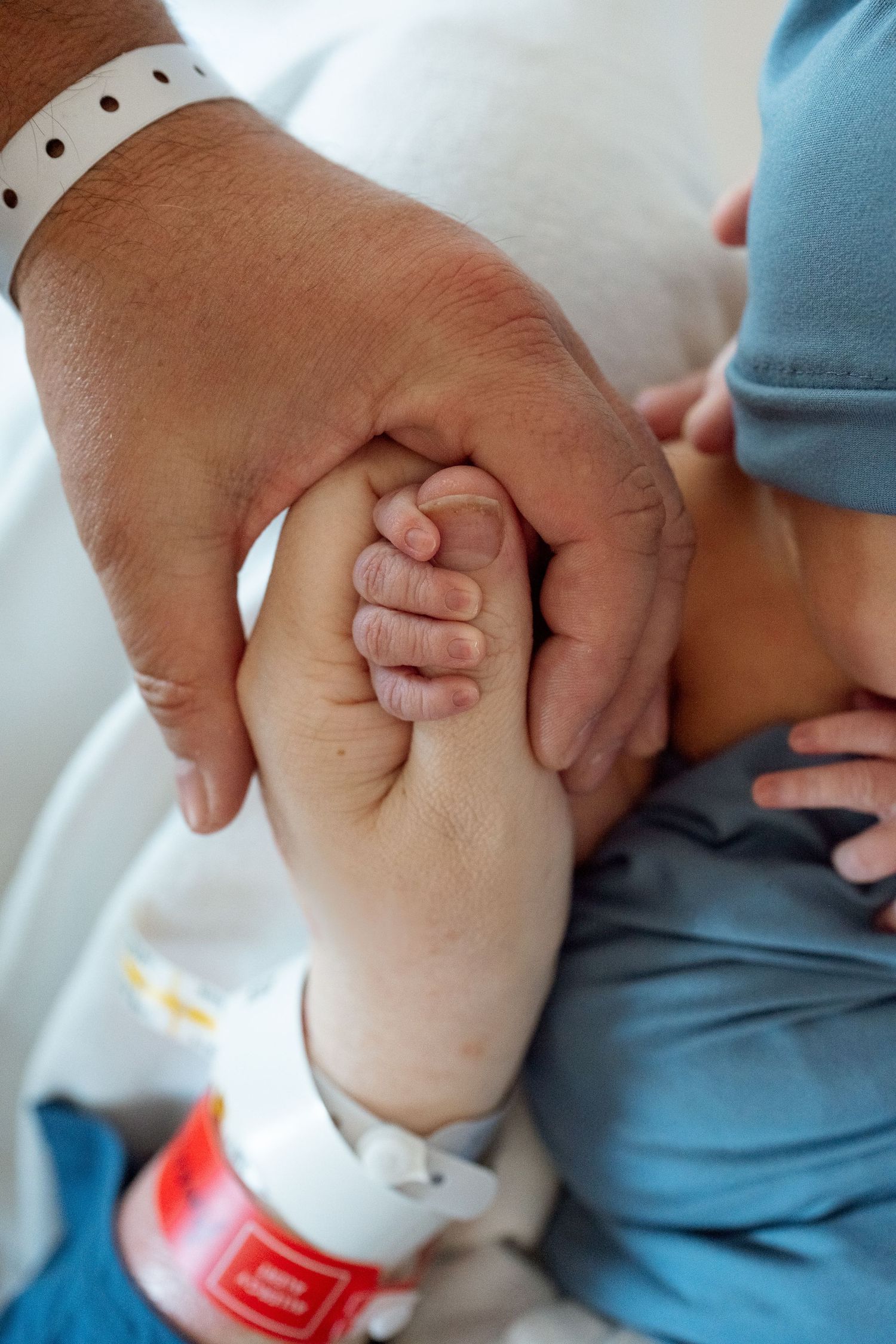 Close up of newborn holding on to mom's thumb, dad's hand wrapped around theirs.  Mom and dad wearing hospital bracelets.