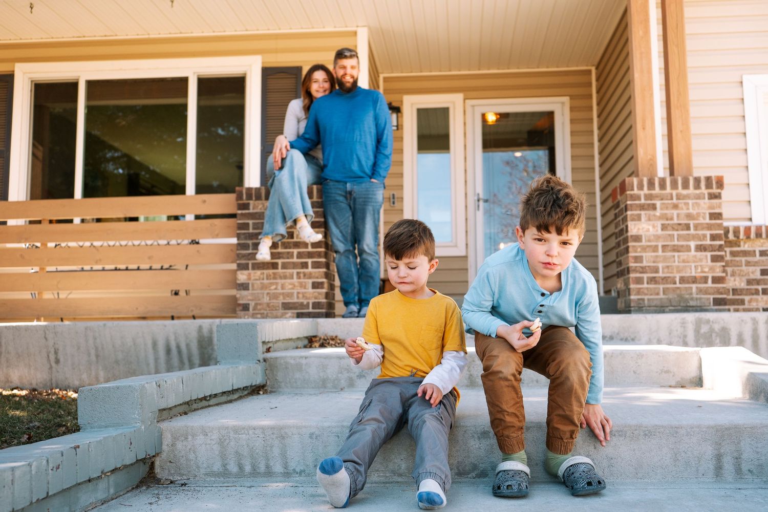 Two boys eating cookies, sitting down on cement steps leading up to house, parents blurry in the background, sitting on their front porch.