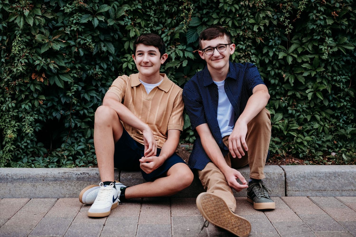 Teen brothers sitting on a curb, green hedge behind them.