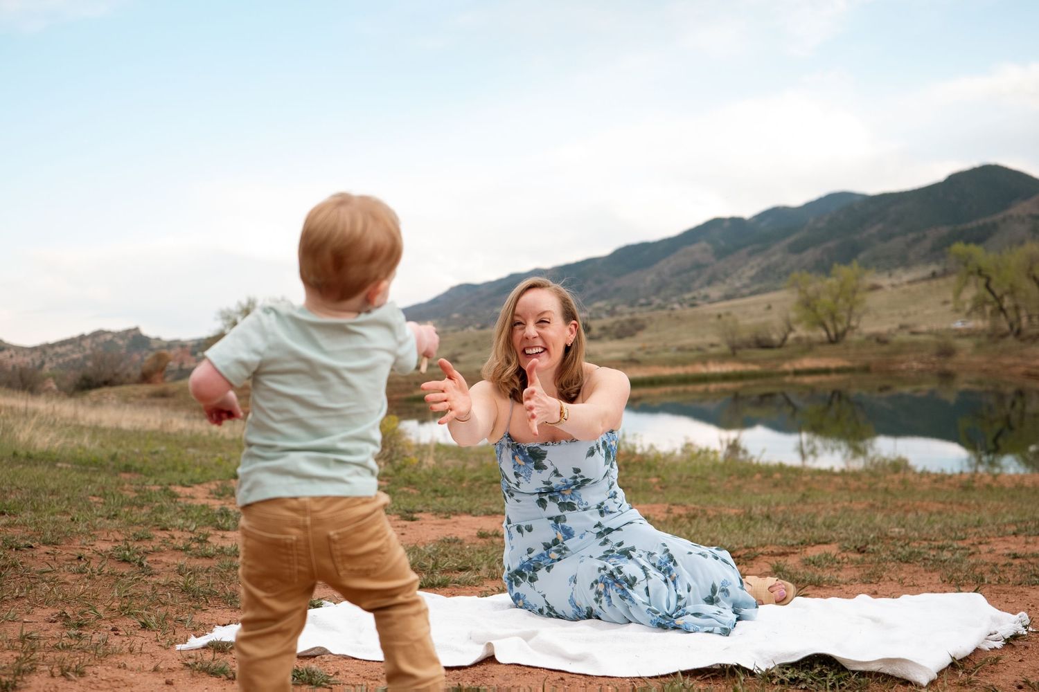 Mother sitting on white blanket holding arms out toward toddler son who is walking toward her, foothills in the background.