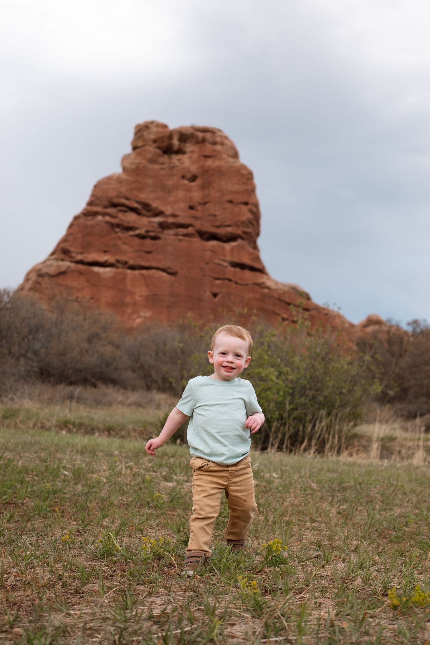 Playful toddler boy walking in field, red rock formation in the background.