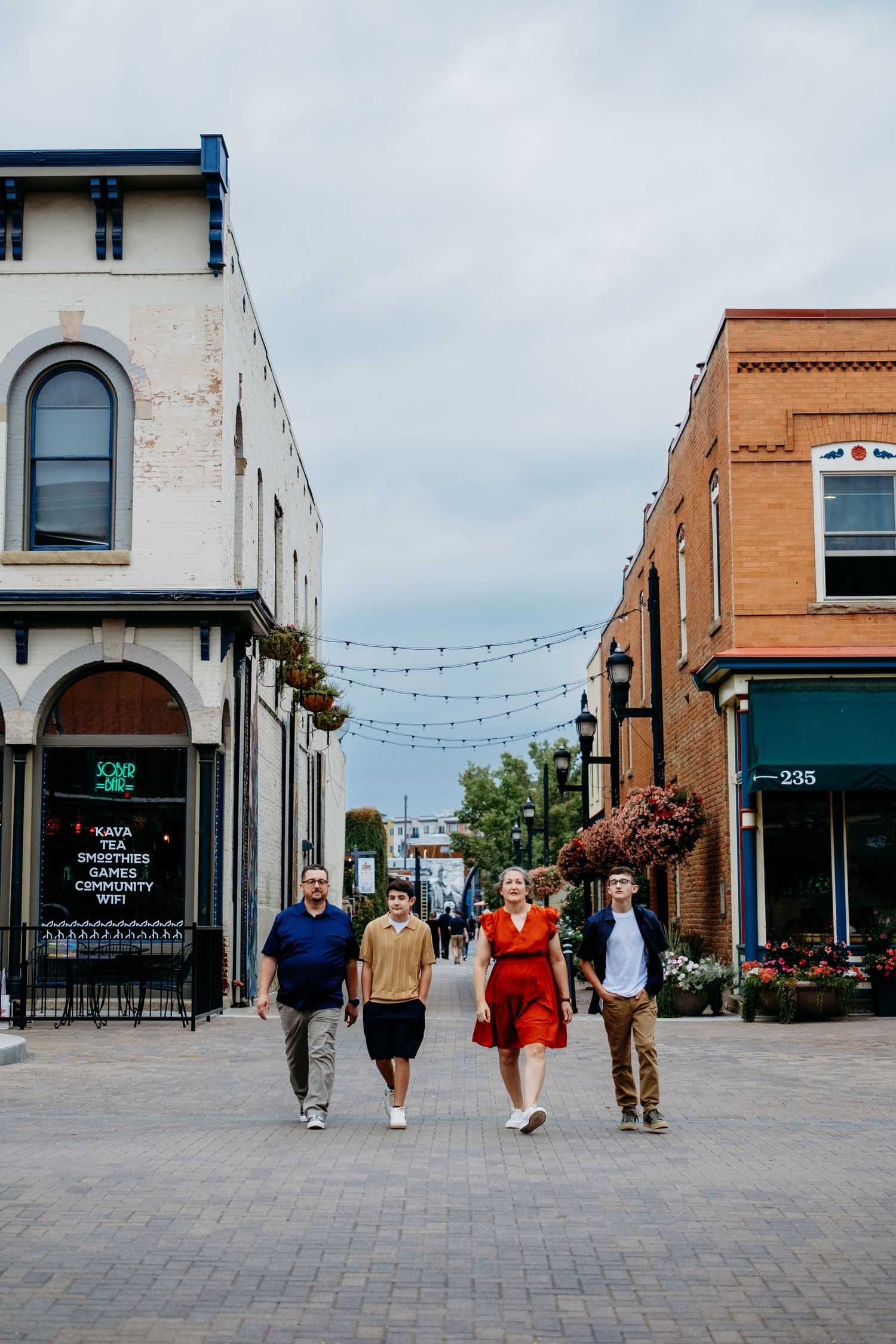 Family of father, son, mother and other son walking across the street in between two beautiful brick buildings, overcast clouds in sky.