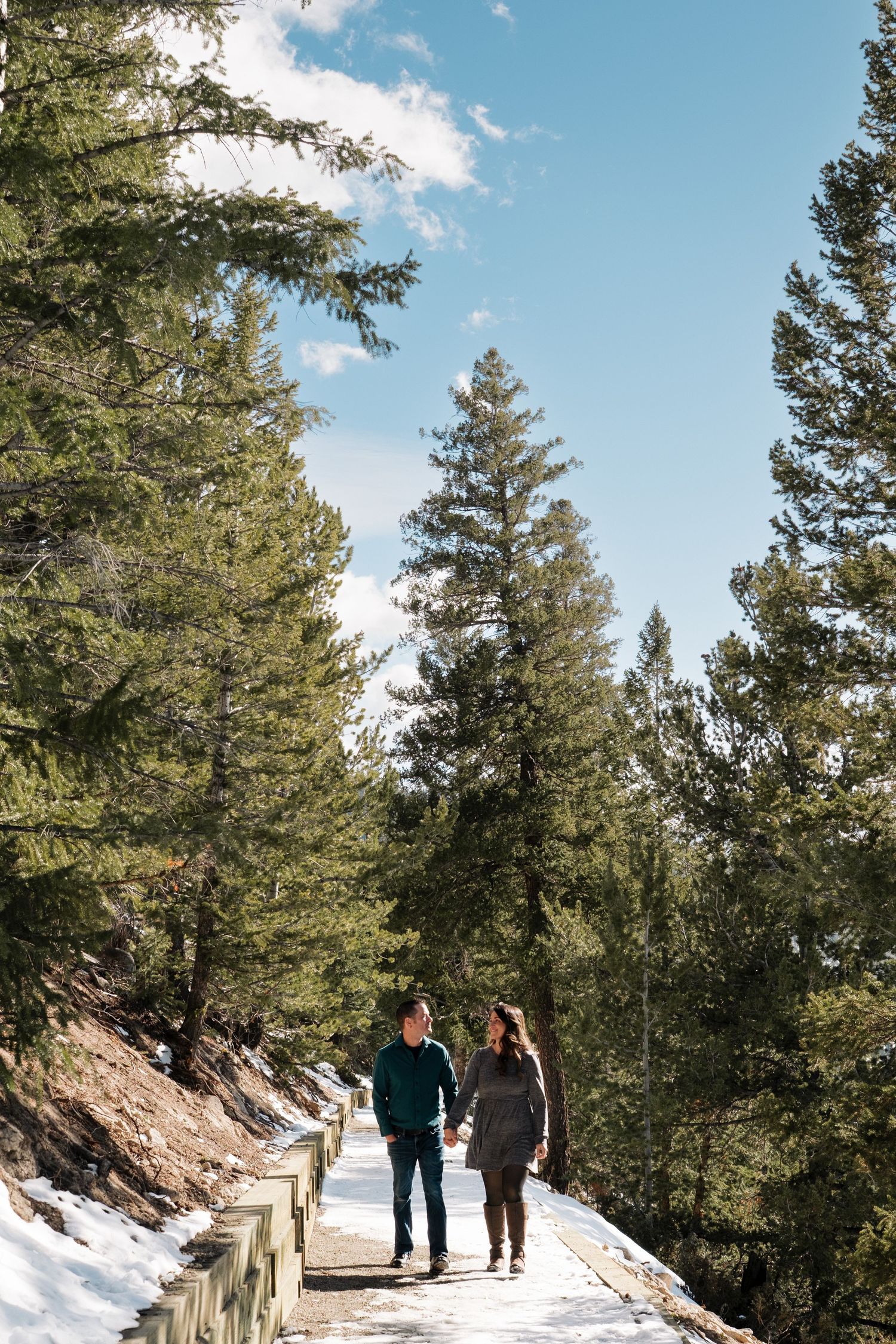 Engaged couple walking together on snowy trail at Sapphire Point Overlook, surrounded by tall trees.