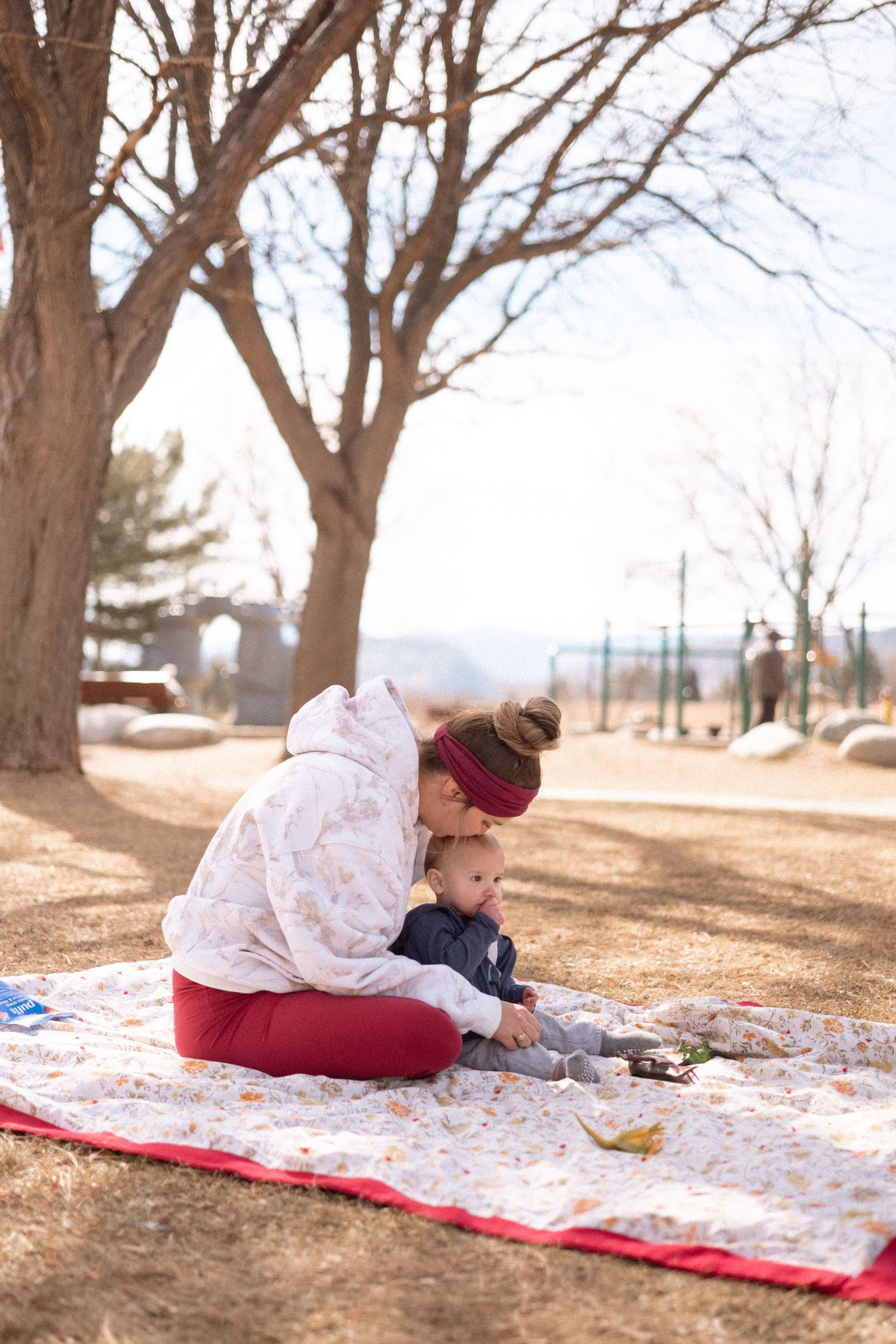 Mother sitting on quilted blanket at the park, one year old son sitting in her lap, playground in background.