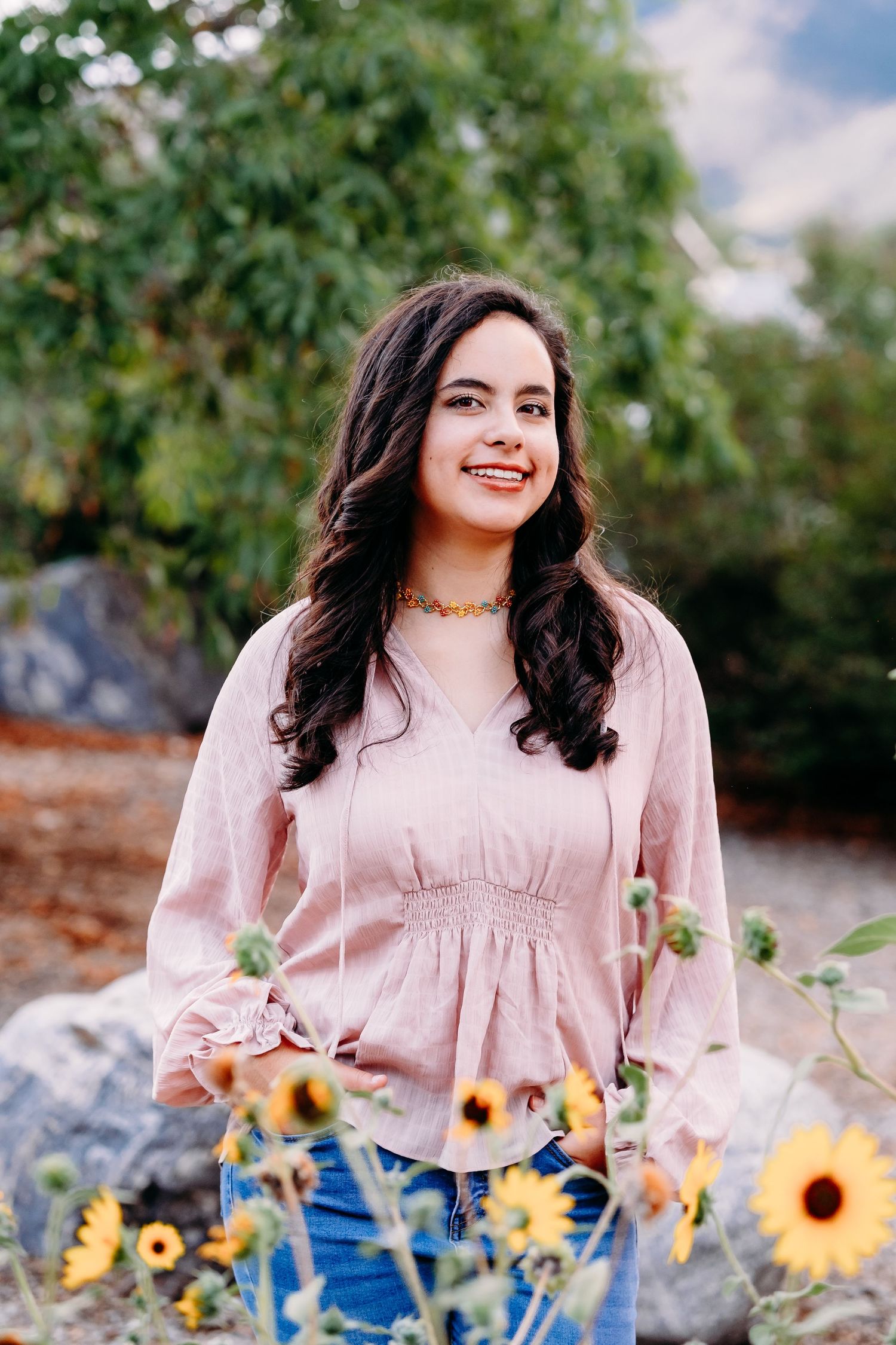 Senior portrait of Littleton graduate, sunflowers in foreground, trees in background, teen smiling.