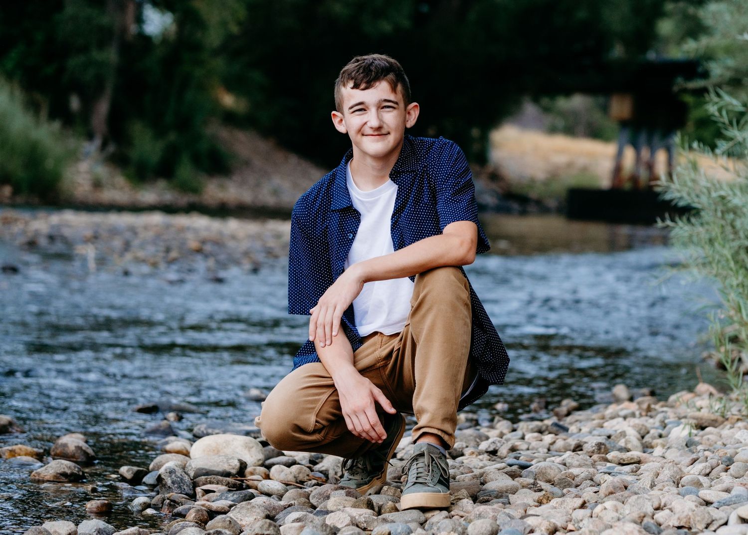 Close up of teen boy, kneeling on rocky bank, near Cache la Poudre river in Fort Collins. Captured by a Denver senior photographer.