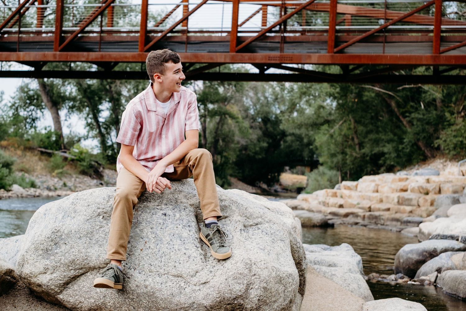 Laid-back senior photo of a teen boy sitting on a large rock by the river in Fort Collins, with a rustic bridge in the background. Perfect e