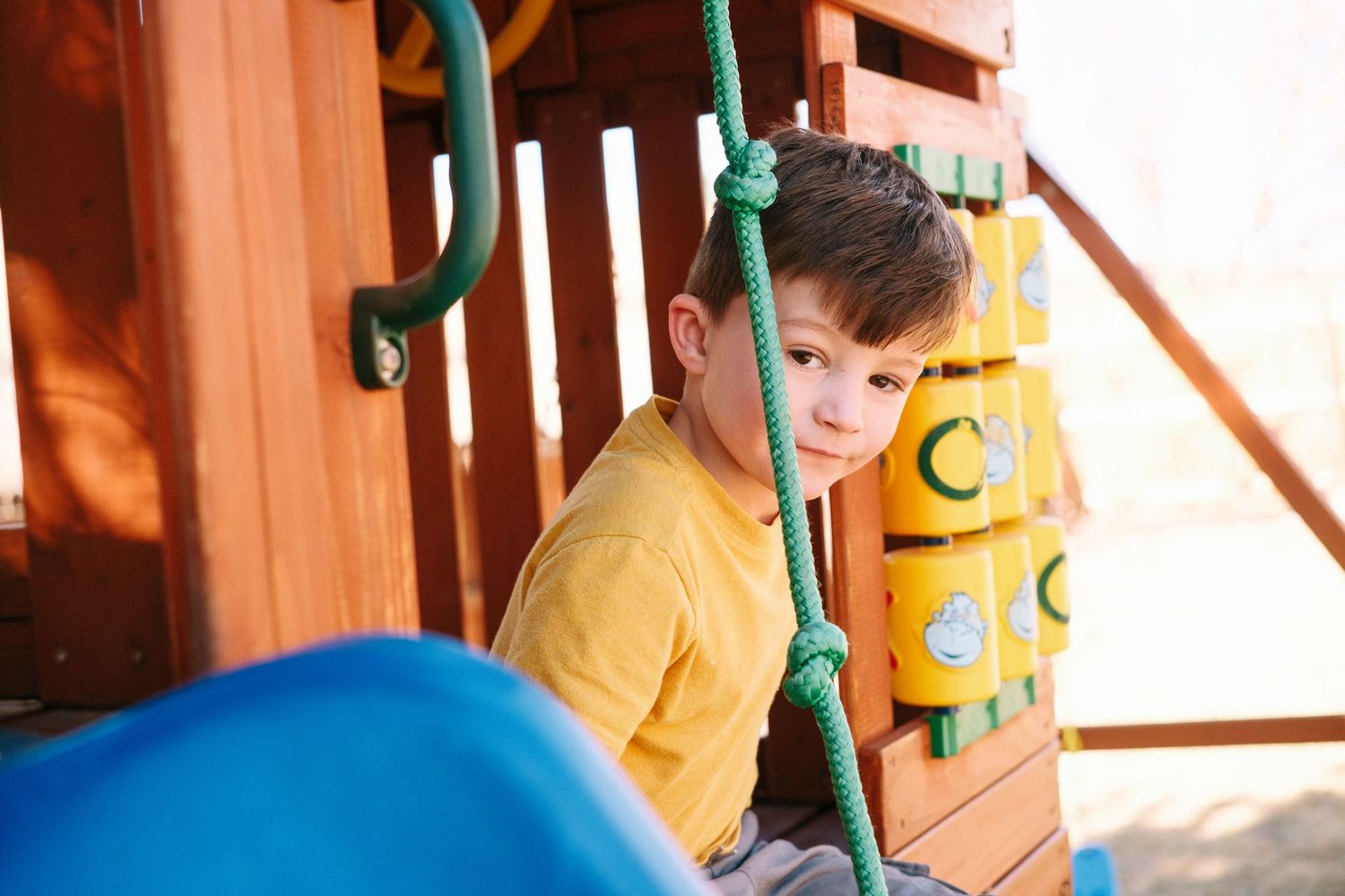 Young boy peaking around climbing rope on backyard playground.