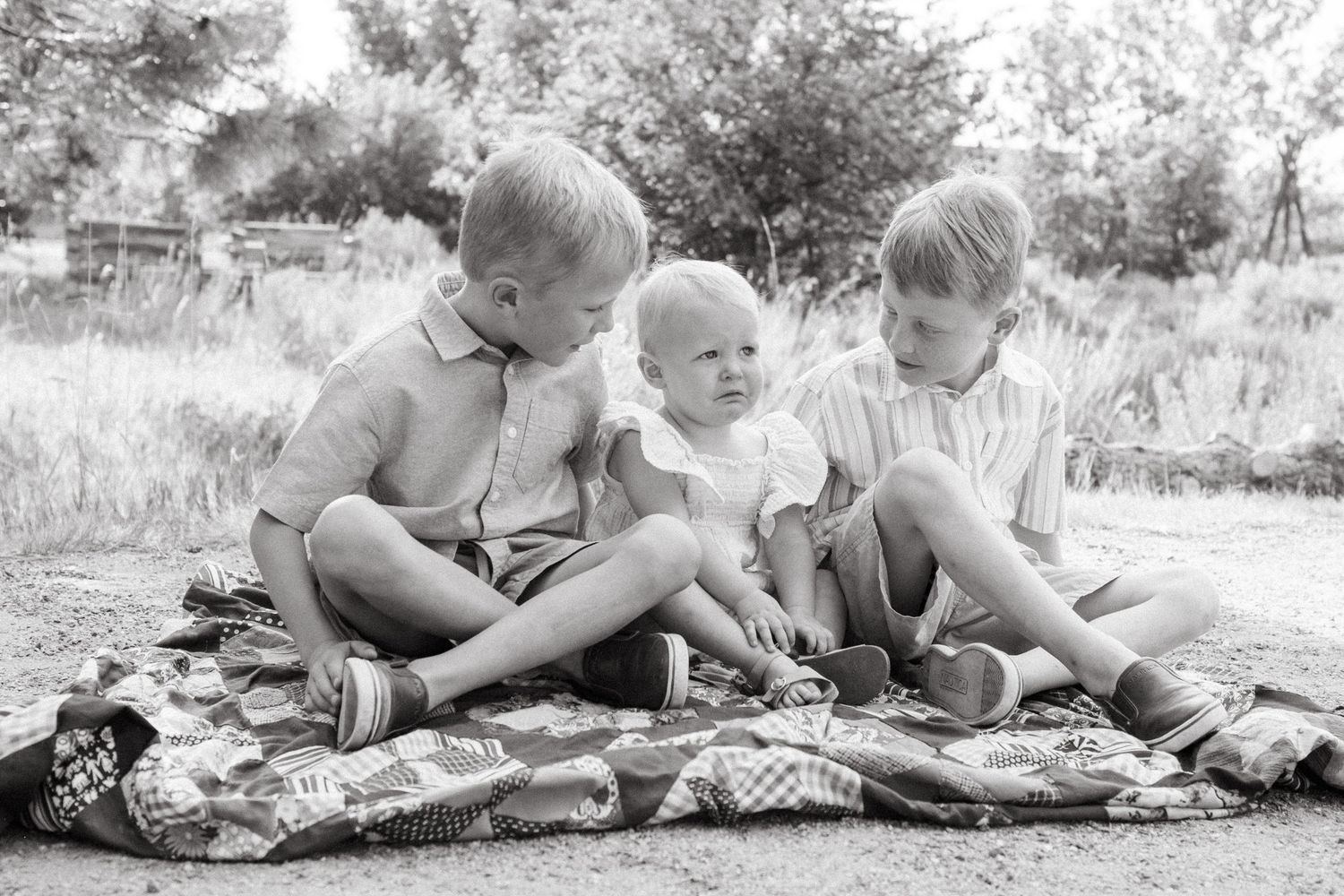 Black and white of two twin brother sitting on each side of their baby sister on a blanket.