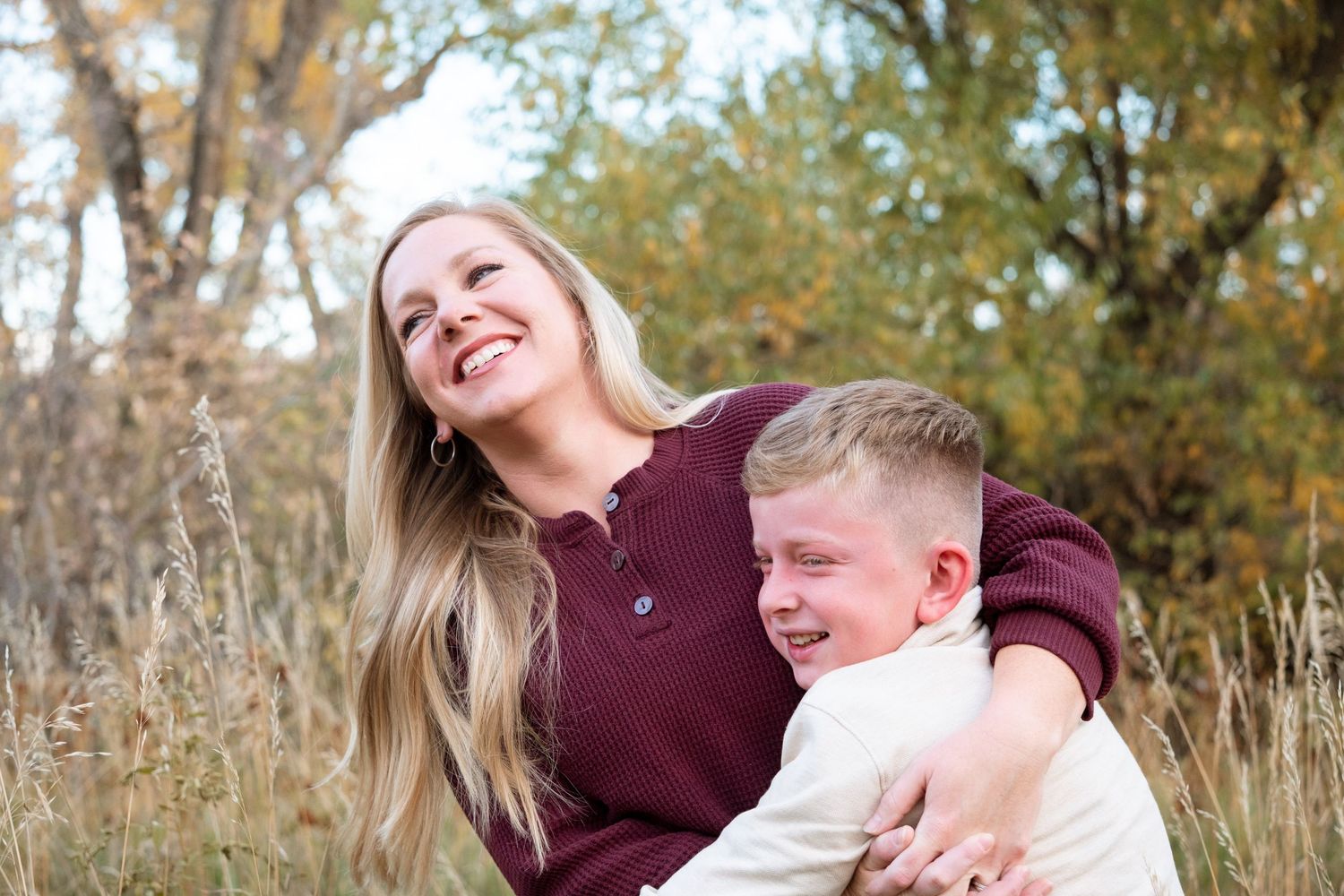 Son playfully squeezing mother, surrounded by tall grasses and autumnal trees in Lakewood, Colorado.