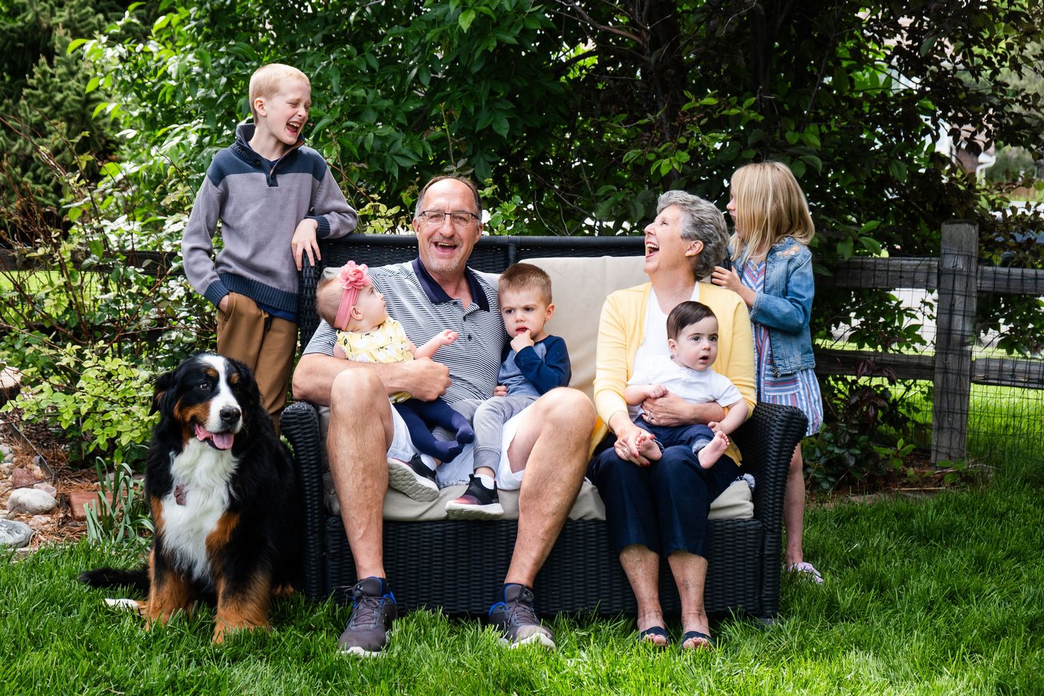 Grandparents sitting on outdoor couch with three grandsons, two granddaughters and Bernese Mountain dog, grandmother laughing.