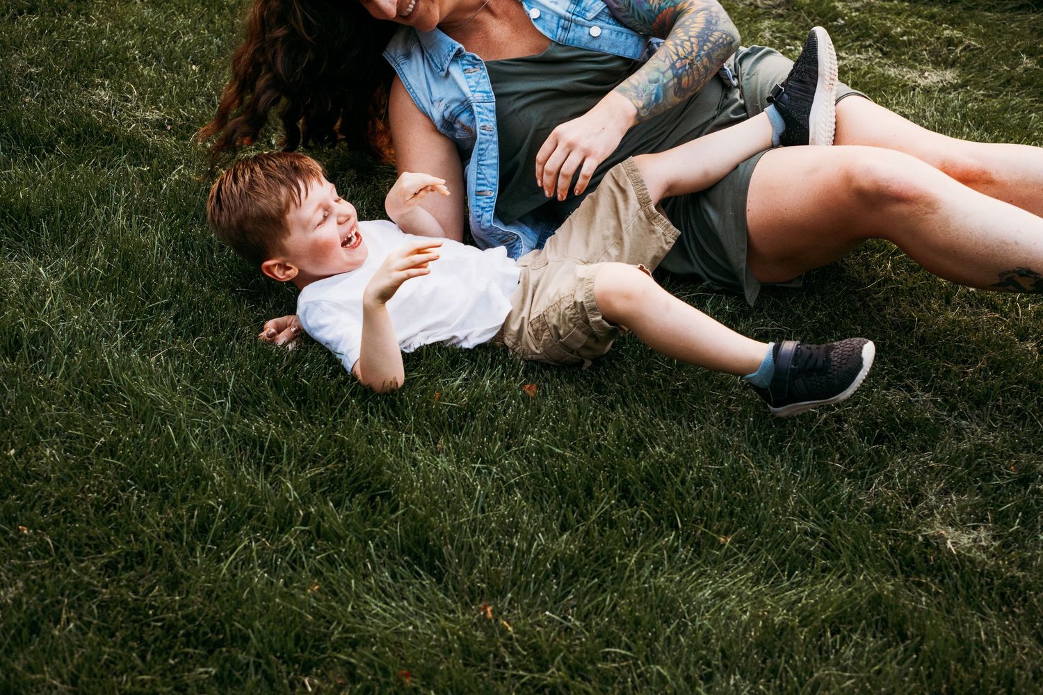 Playful documentary photography motherhood session, mother tickling toddler son on grass.