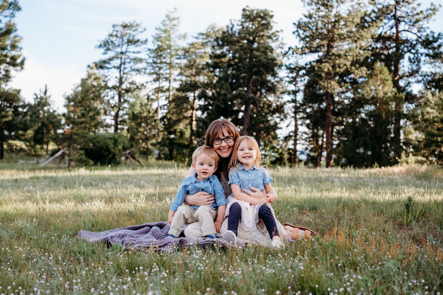 Mother sitting on blanket in field with wildflowers snuggling son and daughter who are sitting on her lap, tall evergreens in background.