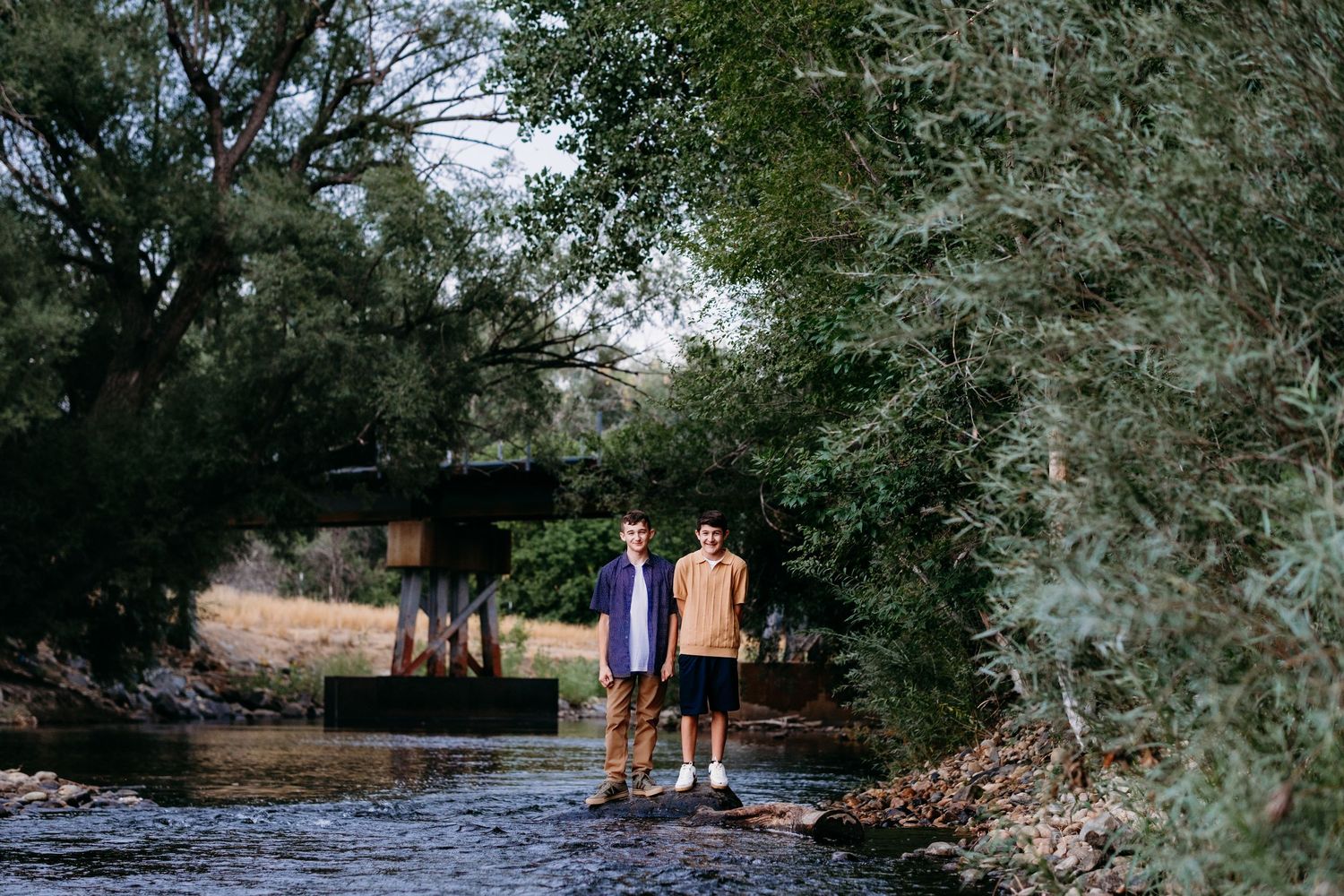 Teen brothers standing on rocks in Cache La Poudre, iron bridge in background, surrounded by lush green trees.