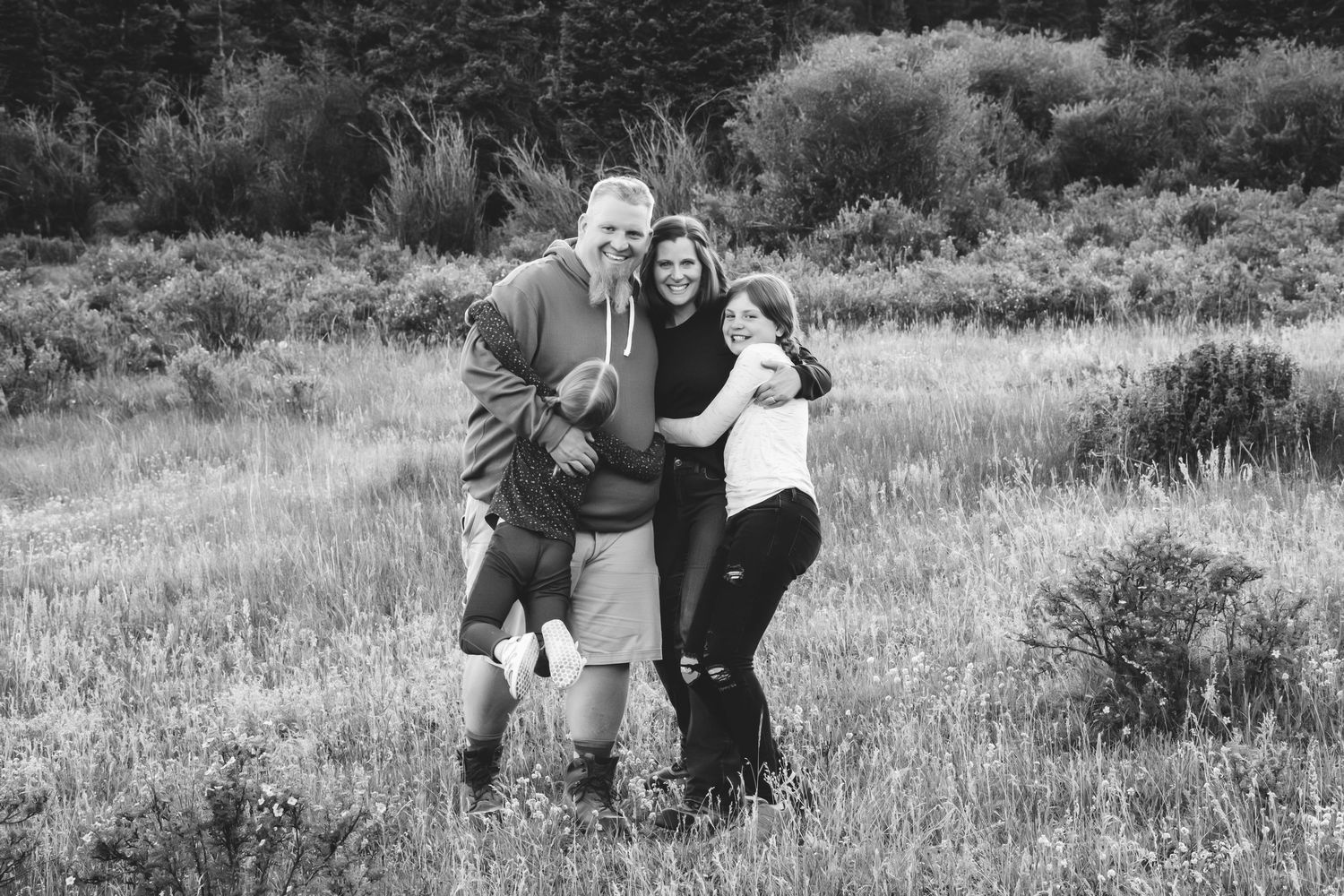 Family of mother, father and two daughters, all hugging closely, smiling, in mountain meadow.