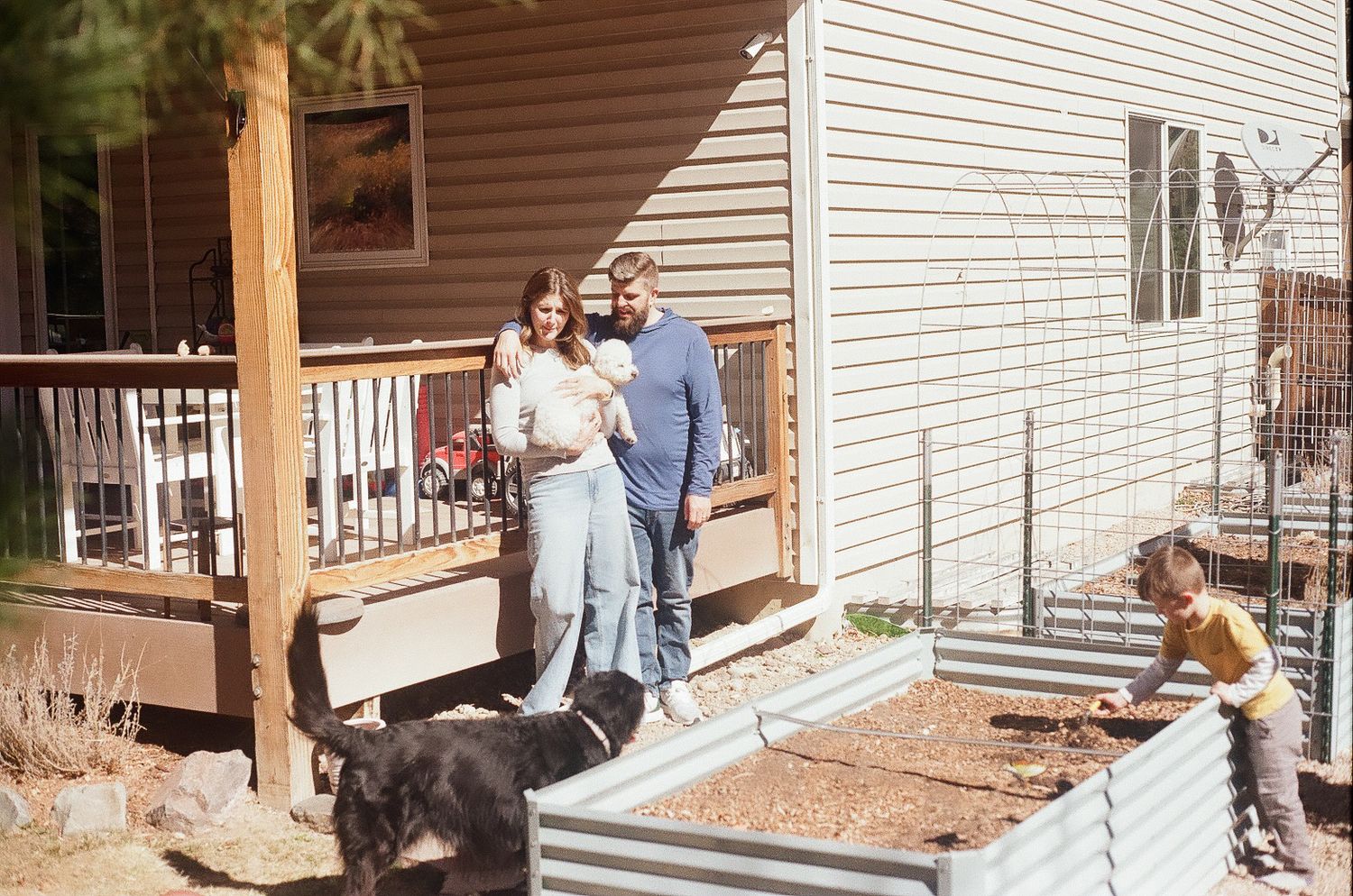 Mother and father standing by the railing of their back porch while kids play in the garden beds.