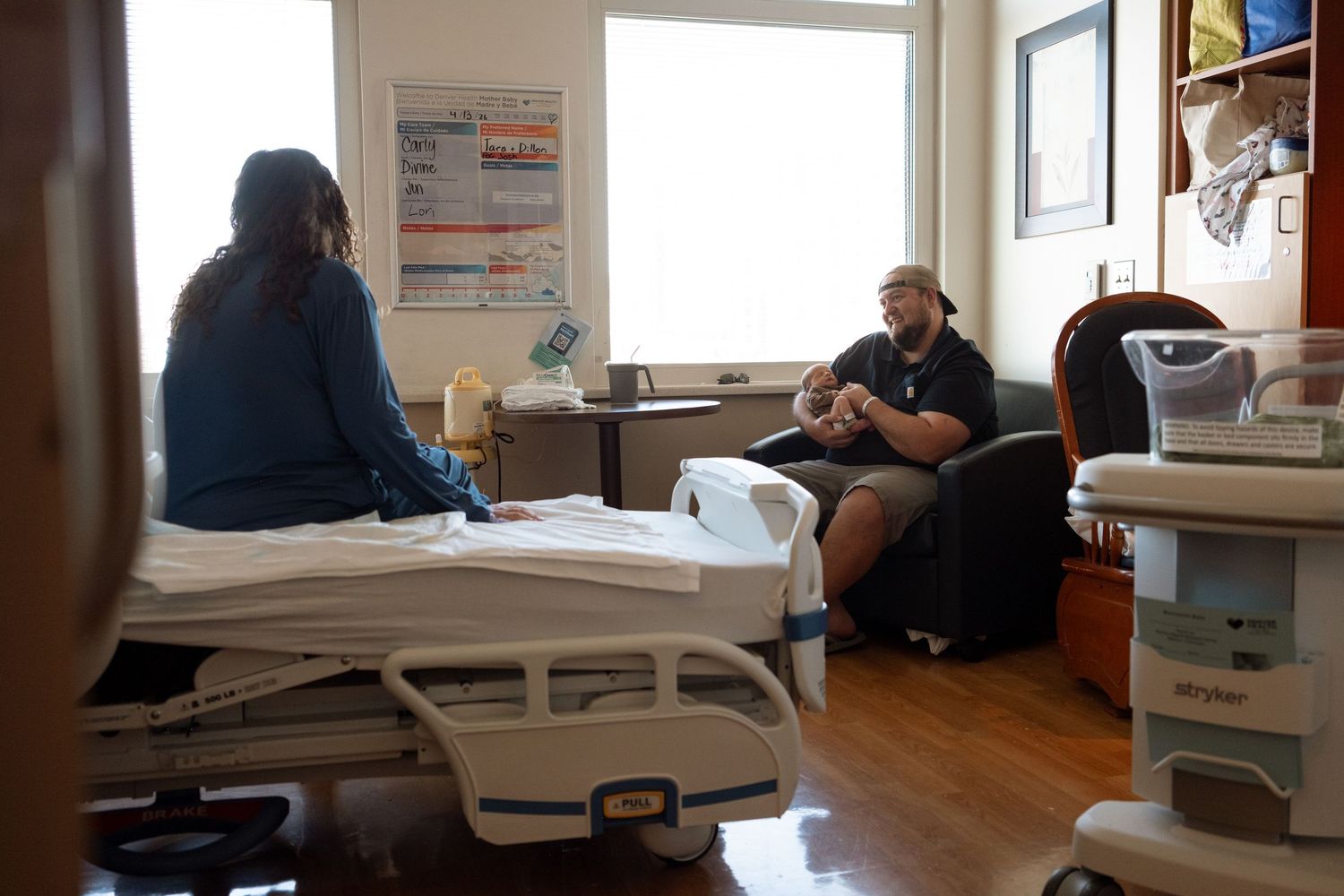 Documentary moment of mother sitting on hospital bed, gazing at father hold newborn sitting on blue chair.