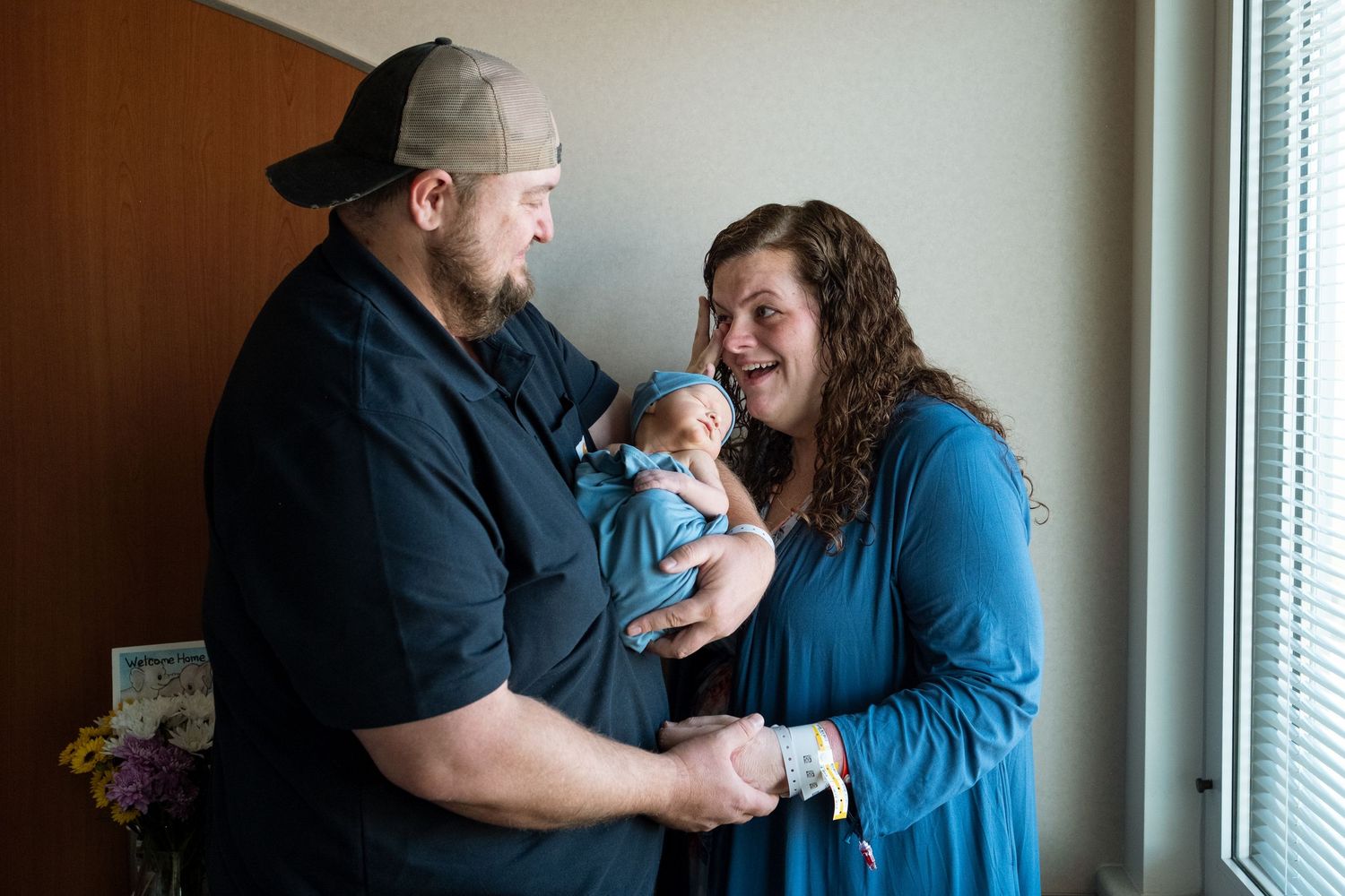 Mother and father standing in hospital room, father holding newborn boy, mother wiping a tear away from her eye.