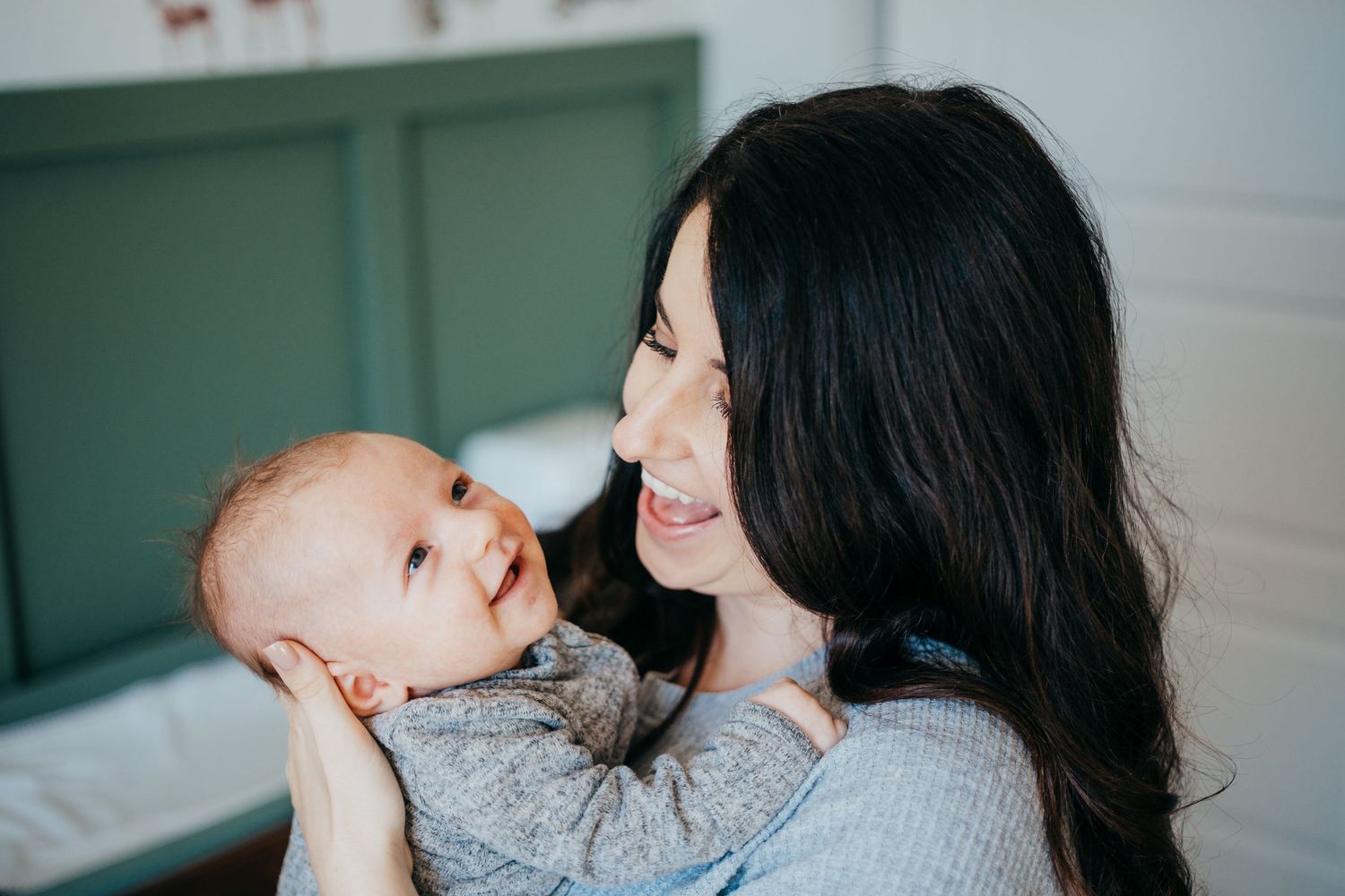 Mother holding newborn baby boy, both smiling and baby gazes toward the ceiling, green nursery walls in background.