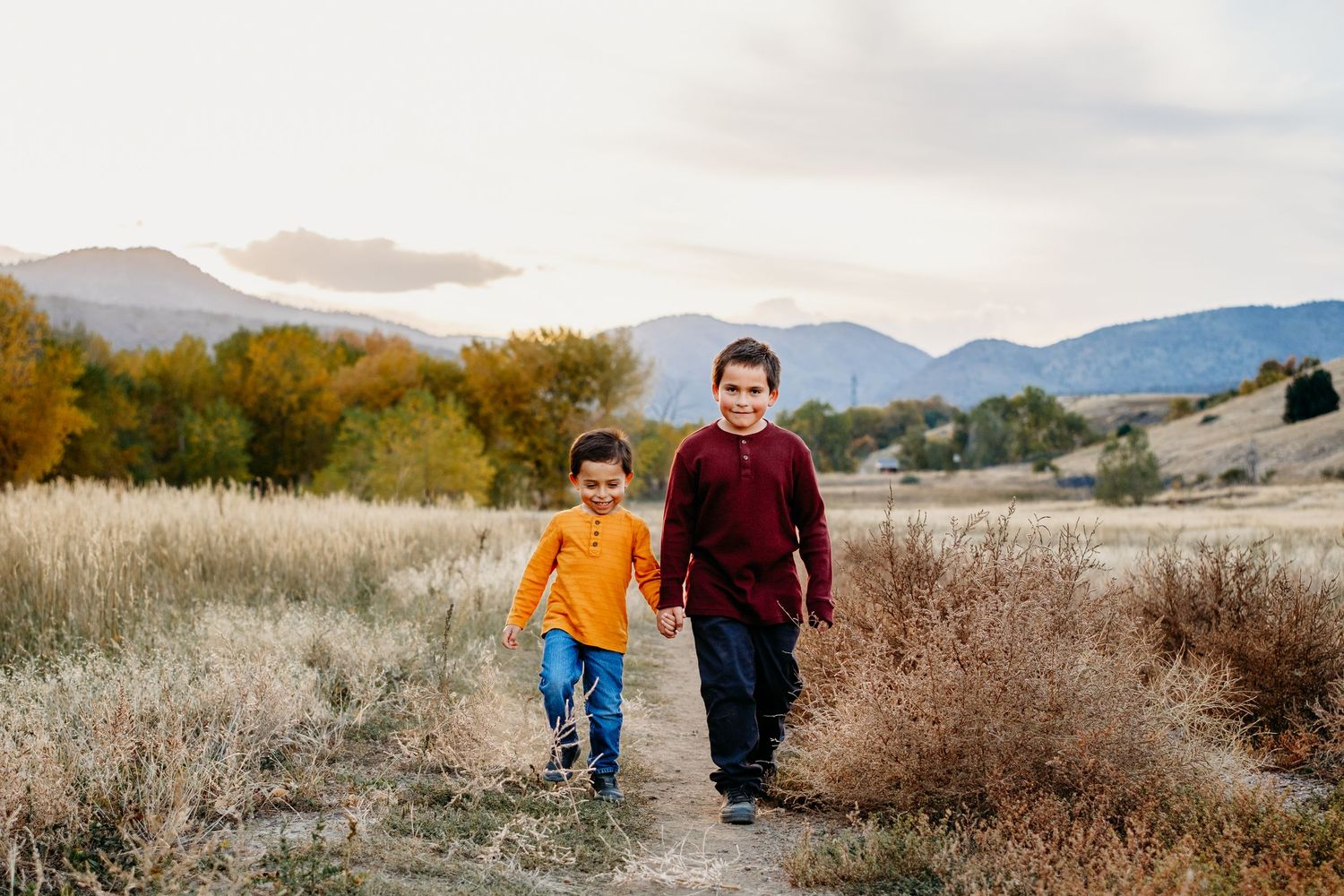 Brothers walking hand in hand in a field at Bear Creek Lake Park, trees and mountain views in the background.