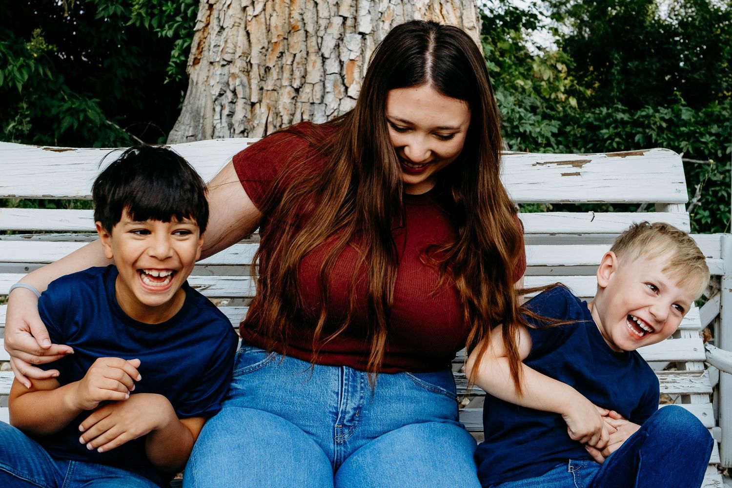 Teenage sister playfully tickling two younger brothers on white swing bench, all laughing.
