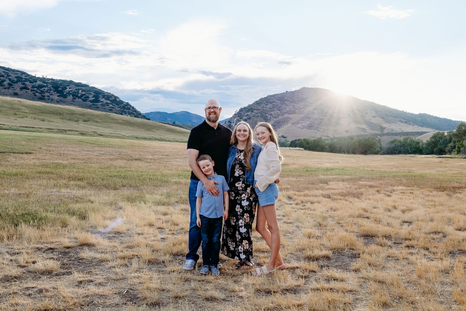 Family of father, mother, daughter and son smiling at the camera in open field with foothills views and sunset in background.