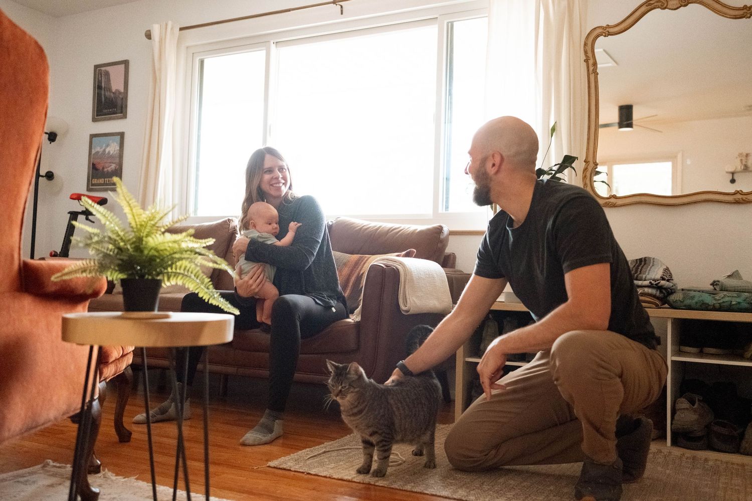 Mother, father, newborn baby and tabby cat all smiling at each other in living room in stunning home session.