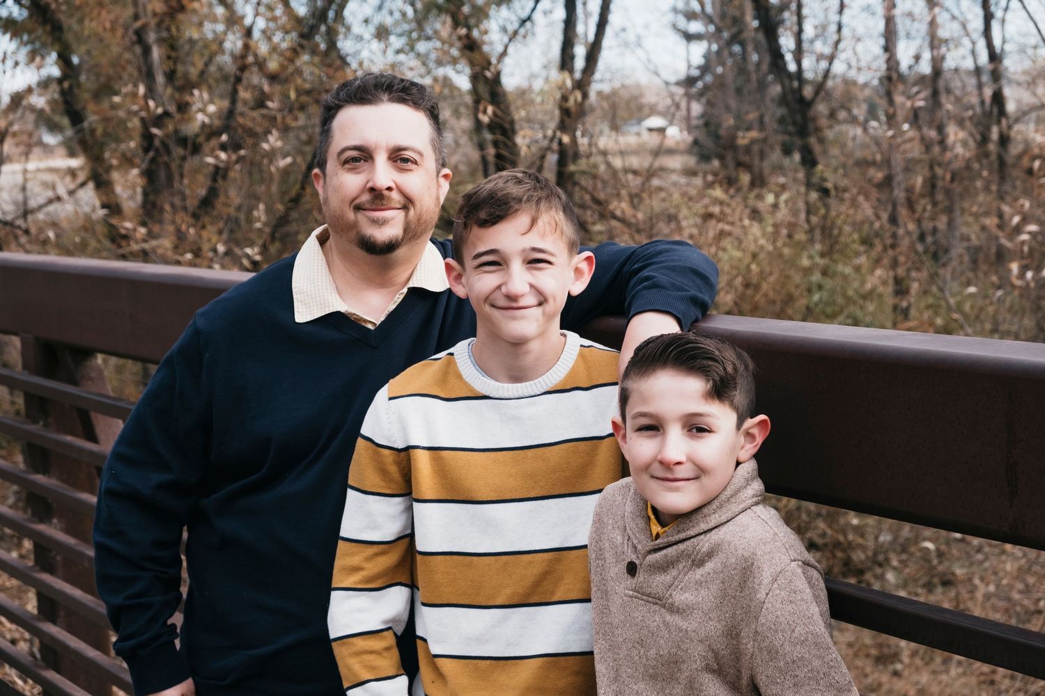 Father and two sons smiling, leaning against rail on bridge.