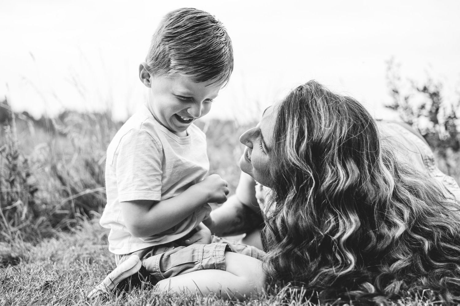 Playful documentary photography motherhood session, child sitting on grass, mother laying down smiling up toward him.