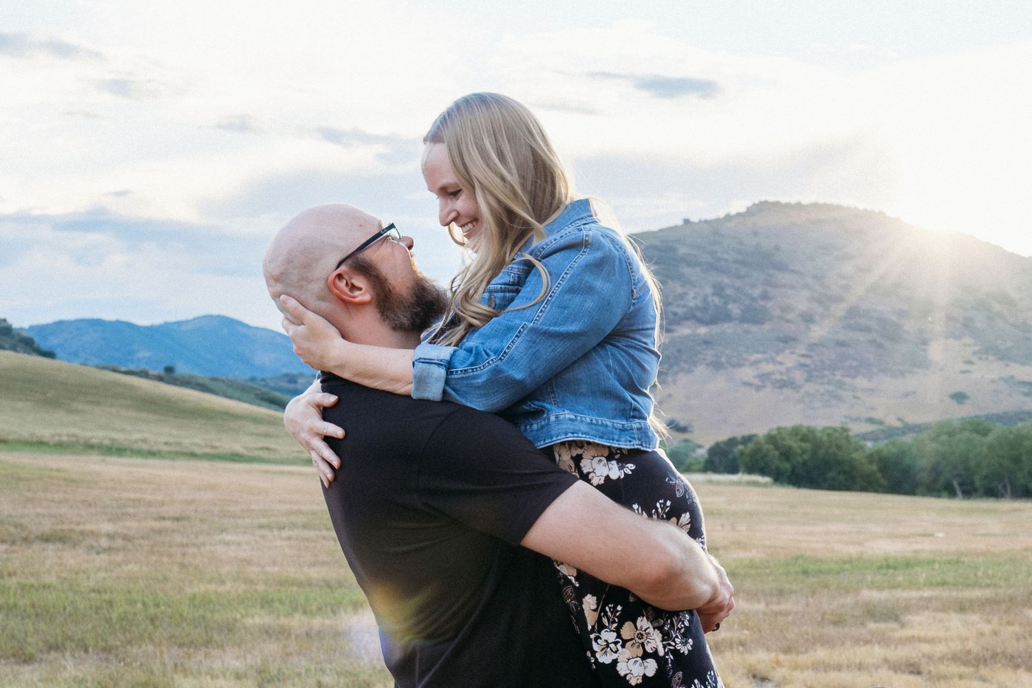 Husband and wife embracing in field, husband lifting wife off the ground, sunburst peaking over foothills in background.