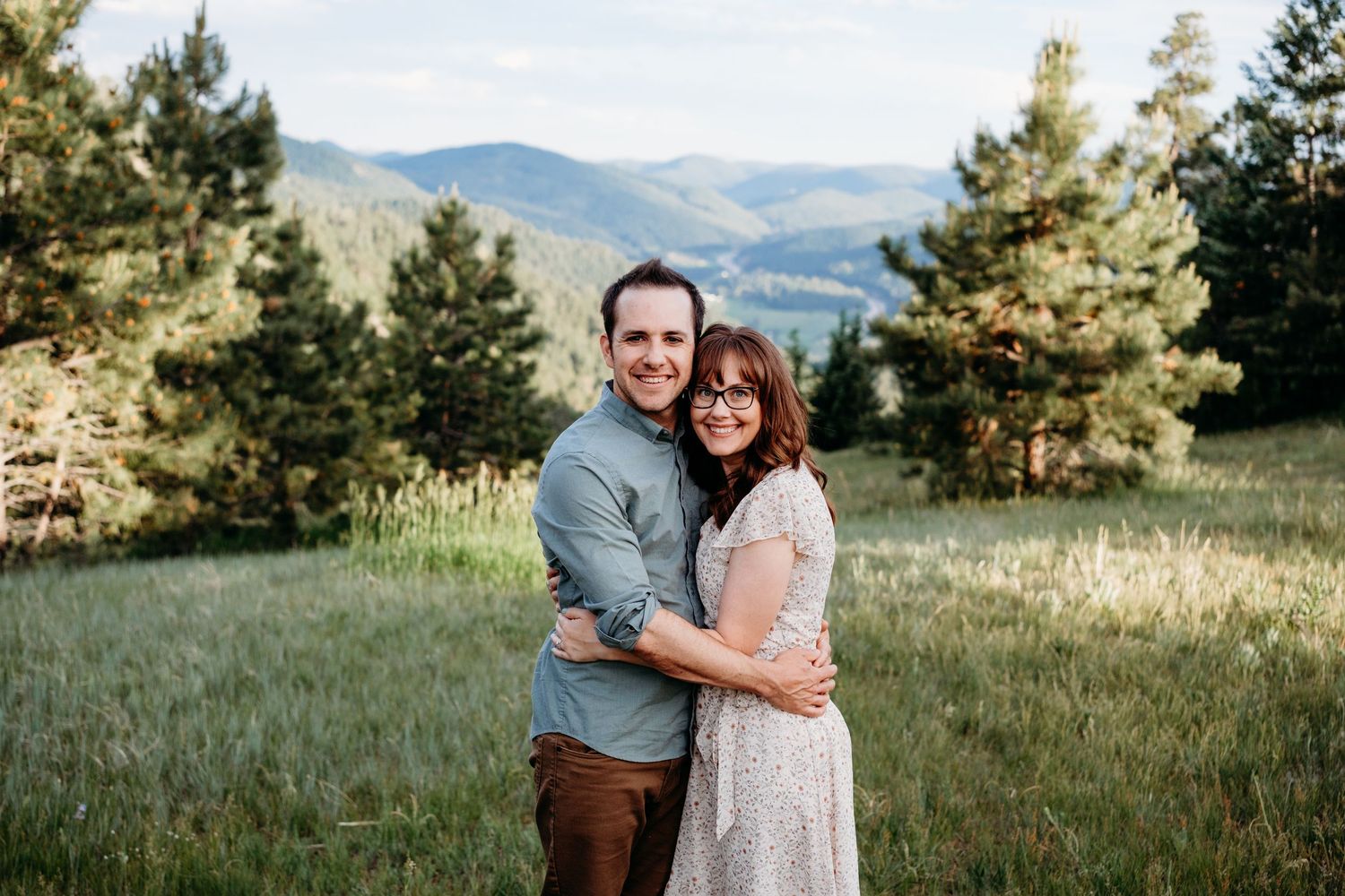 Adult couple standing close together, belly-to-belly, embracing one another, forest and mountain views in background.