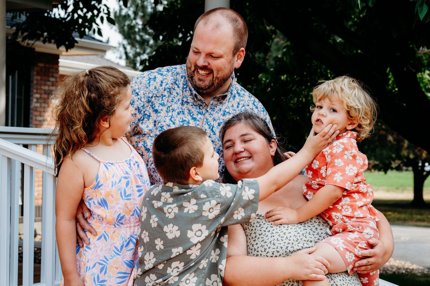 Close up of family of five standing together, all looking at one another, brother playfully squishing younger brother's cheeks.