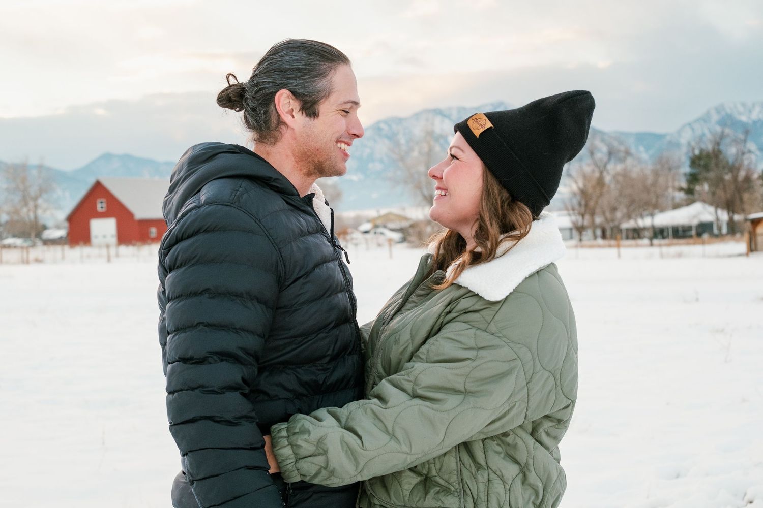 Man and woman smiling toward one another on snowy day, snow capped flat irons and red barn in the background.