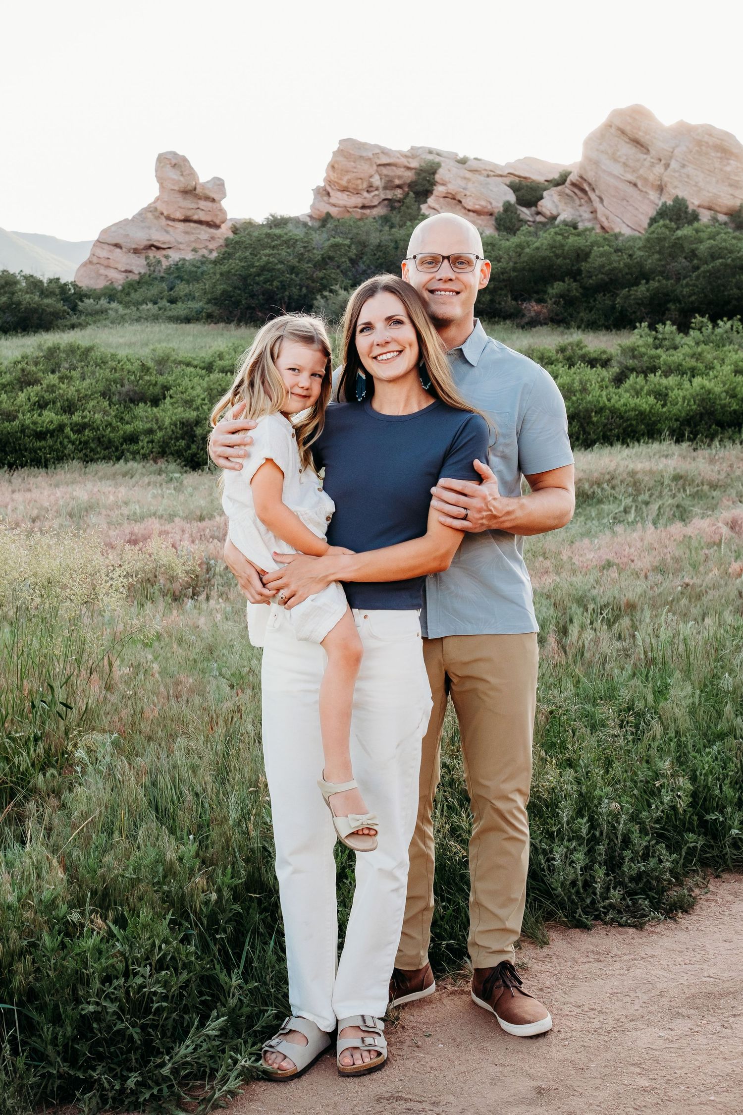 Family of three - mother holding daughter, daughter tightly holding on to mother, father standing next to them, alls miling.