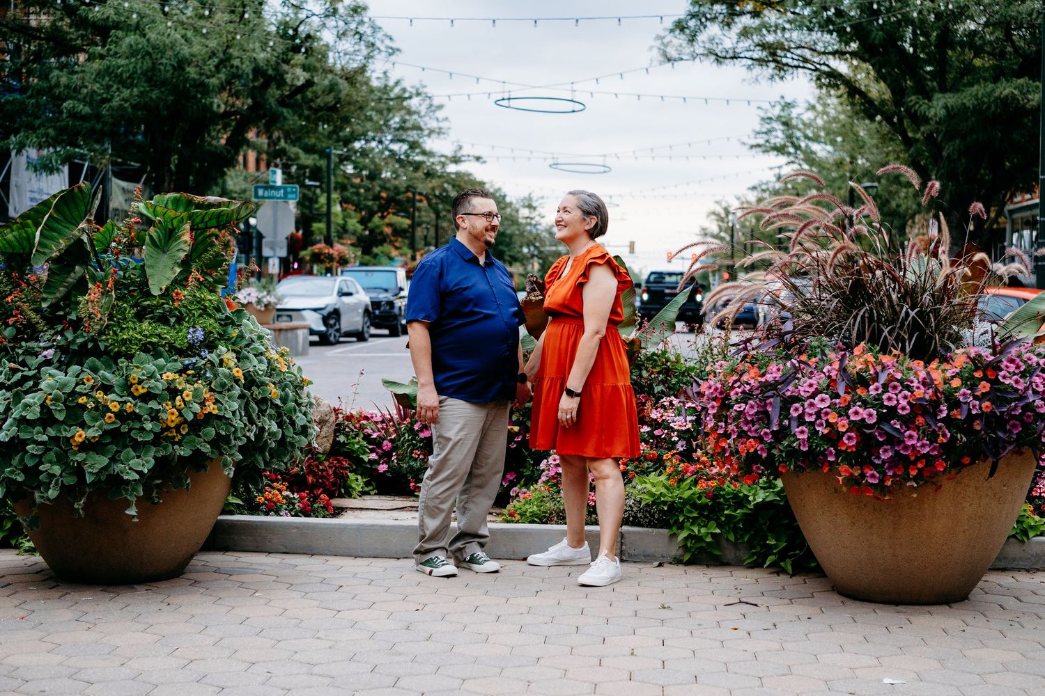 Adult couple standing together in downtown Fort Collins, smiling toward one another, surrounded by lush planters overflowing with flowers.