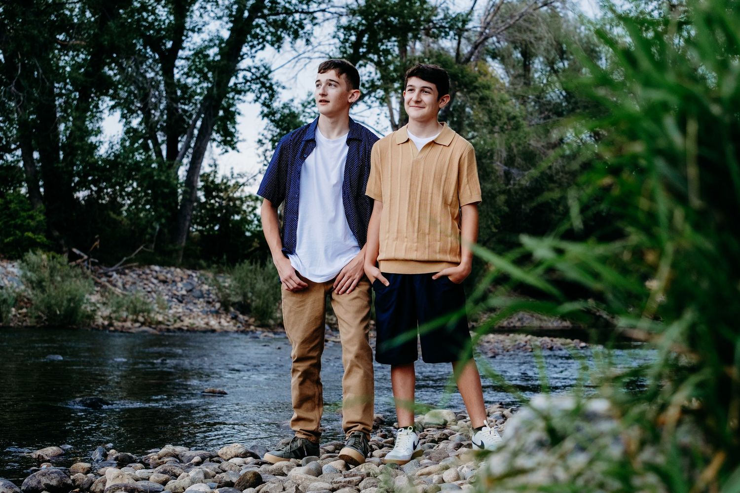 Brothers standing together on rocky bank at Cache la Poudre in Fort collins, surrounded by lush greenery of trees and shrubs.