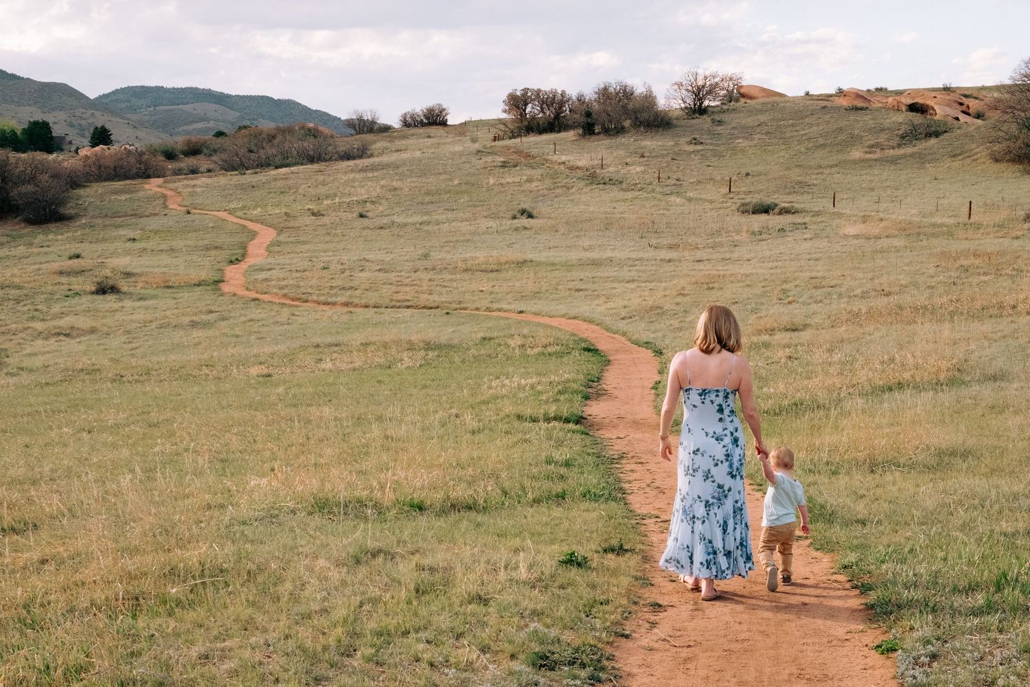 Mother and toddler son walking on a meandering, dirt trailhead, stunning landscape in the background.