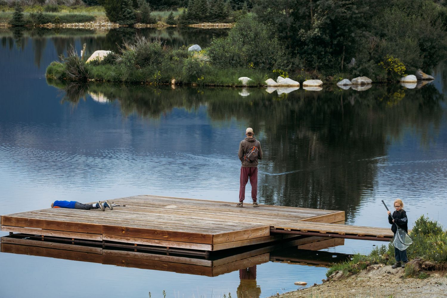 Father fishing off dock with son laying flat on dock peering into pond, daughter on land with a fishing net.