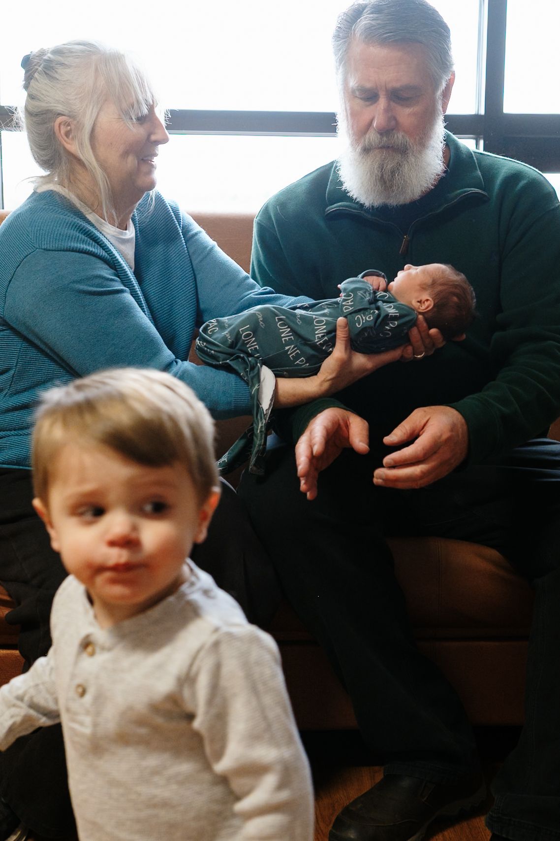 Grandmother handing newborn grandson to grandfather, toddler grandson walking in foreground.