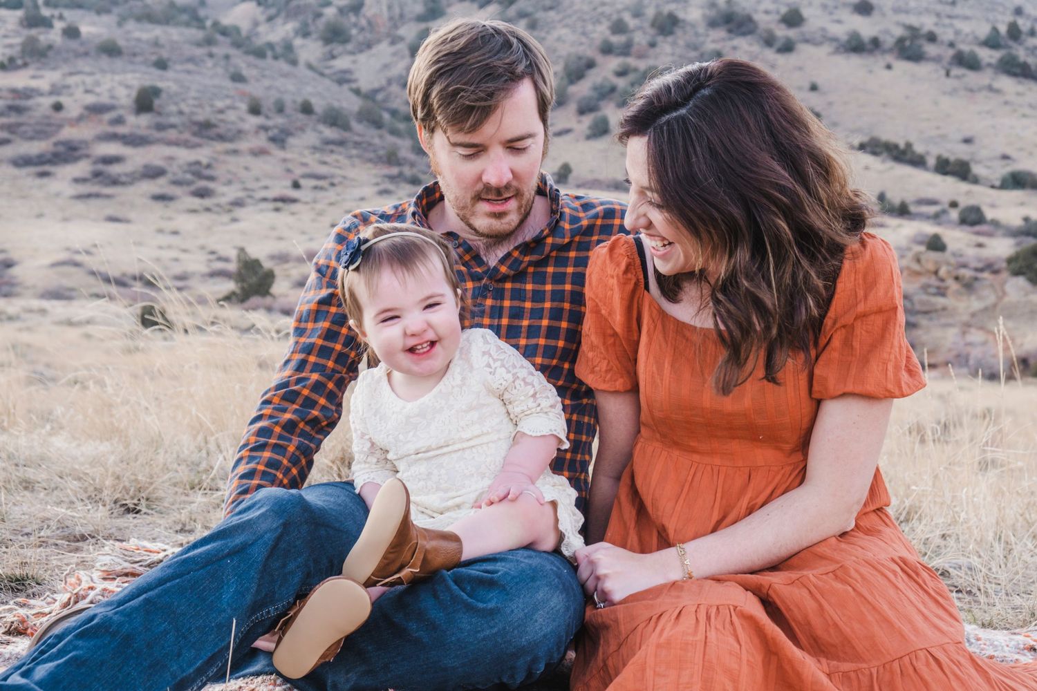 Mother, father and toddler daughter sitting close together at Mount Falcon East in Morrison, Colorado, daughter laughing.