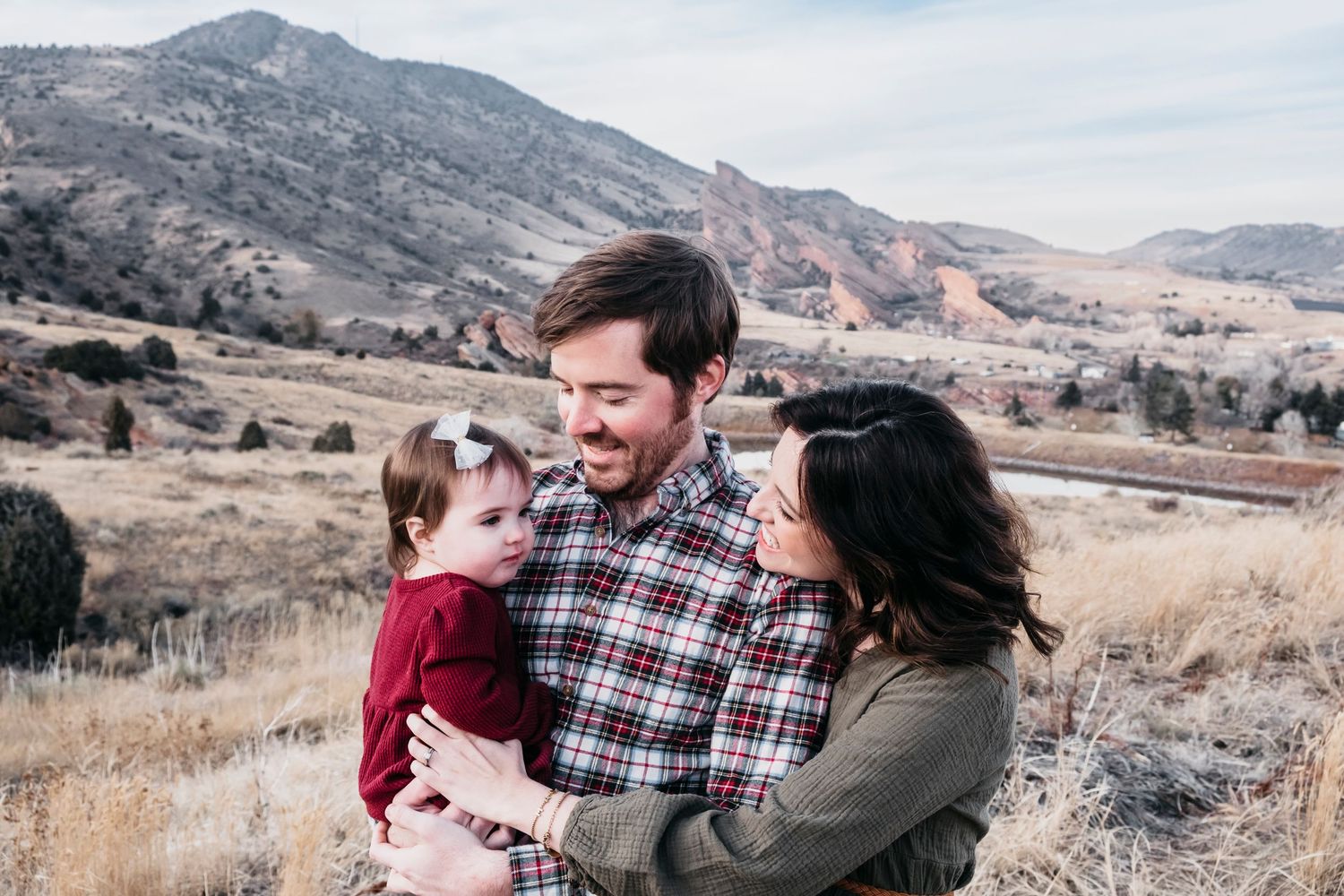 Mother, father and toddler daughter snuggling close together at Mount Falcon East in Morrison, Colorado, foothills and Red Rocks in the back