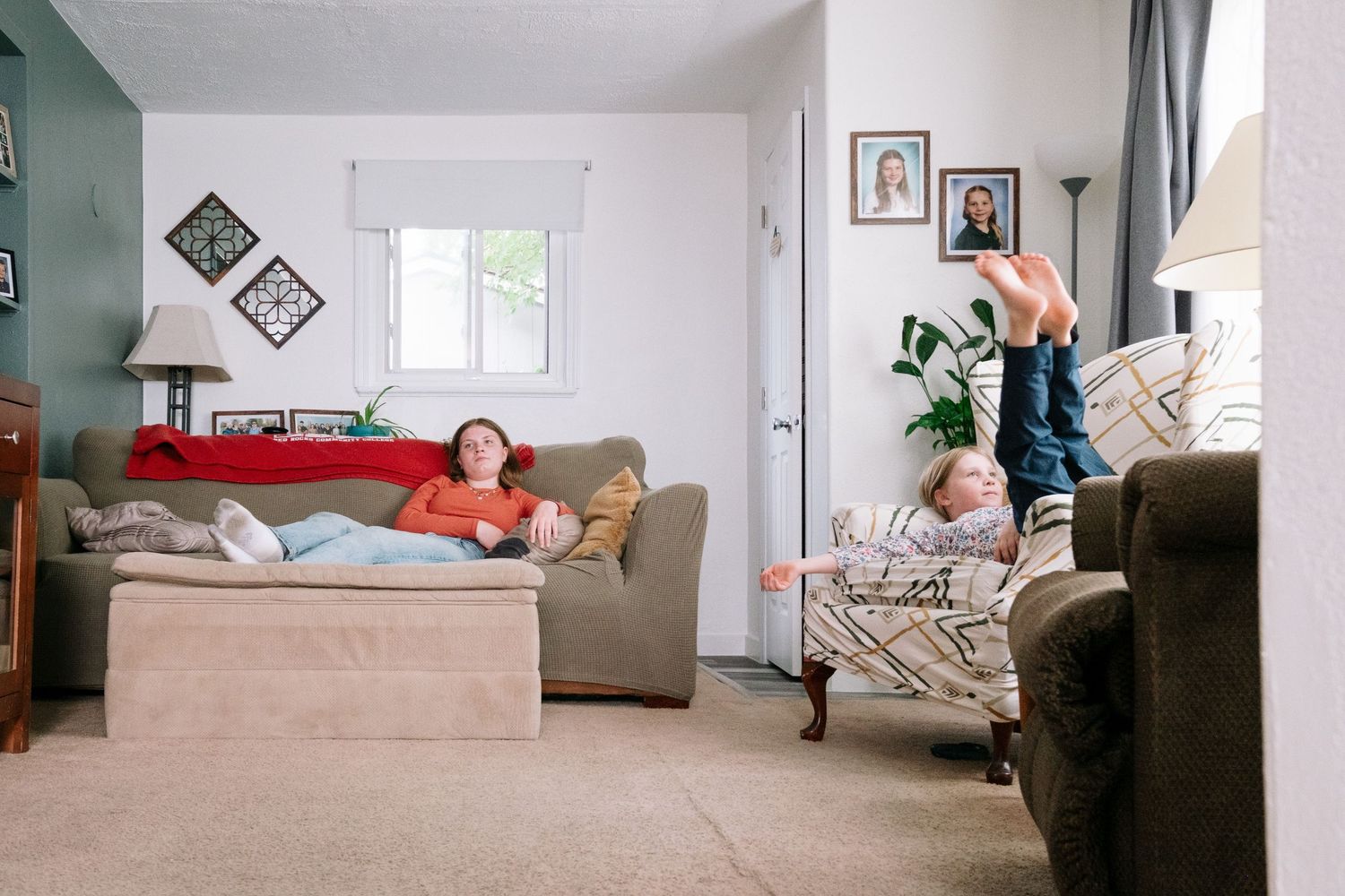 Playful documentary portrait of two sisters sitting on two different couches in living room, one with feet up in the air.