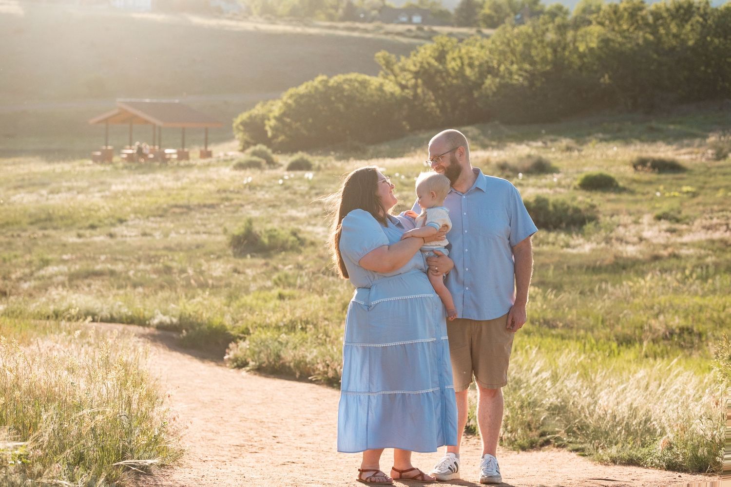 Father and mother standing close together, mother holding son, grasses, shrubs and wooden picnic area in the background.