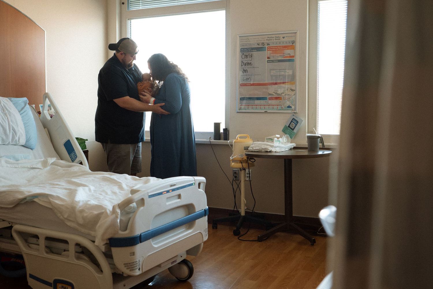 Mother and father standing by the window in their hospital room.  Father is holding baby while mother kisses the baby's forehead.